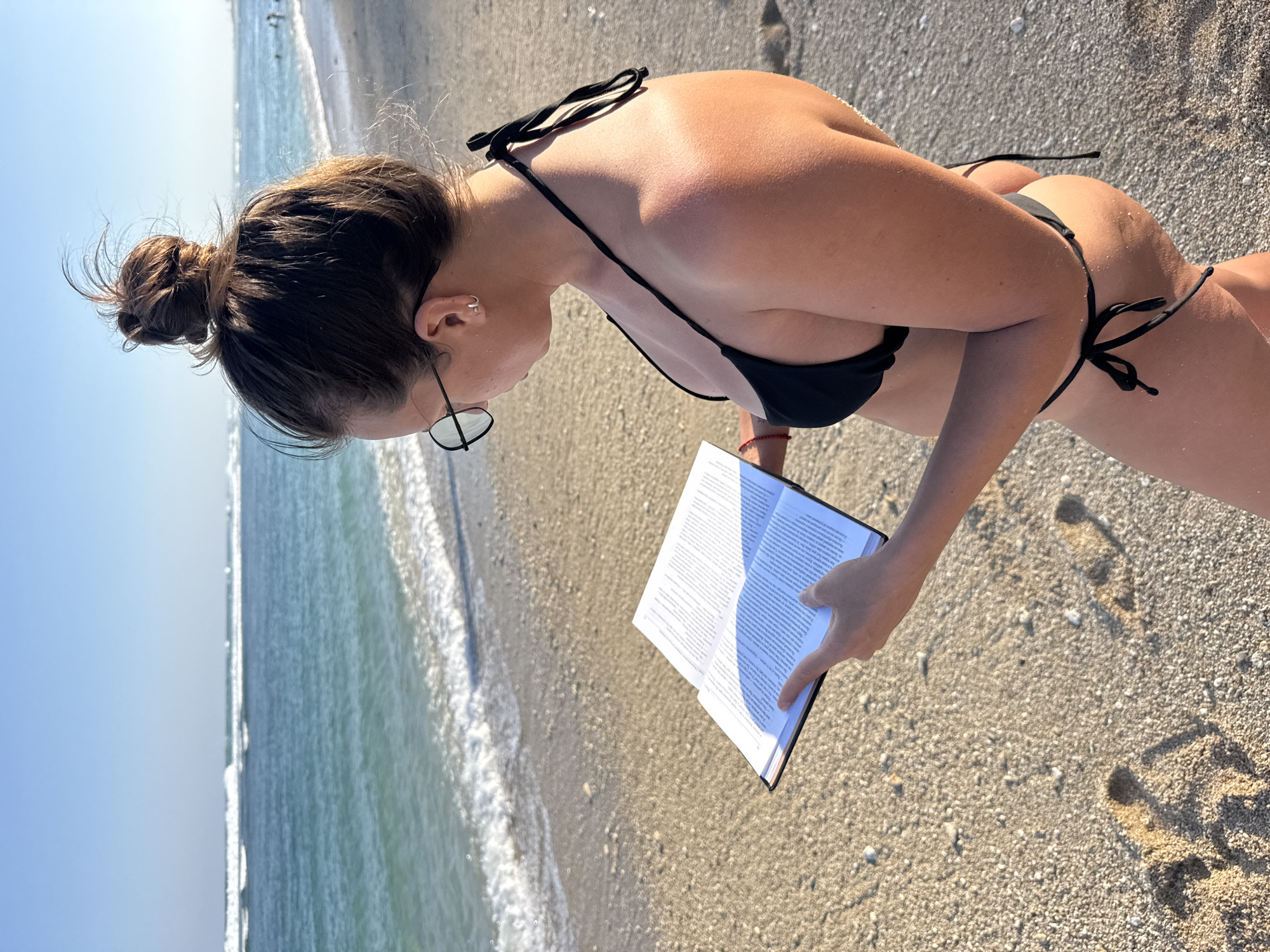 A high-angle, eye-level outdoor shot captures a solitary female reading a book on a sunlit beach in Kuta Selatan, Indonesia. The woman, viewed from slightly behind her right side, has brown hair styled in a messy bun and wears black, round-rimmed glasses. She is dressed in a black string bikini, revealing tanned skin. Her gaze is directed downward, focused on an open book with white pages densely filled with text, held in her left hand. The beach foreground consists of light brown sand interspersed with small pebbles, bearing visible footprints. Gentle waves with white foam break along the turquoise ocean shoreline, which stretches into deeper blue waters under a clear, bright blue sky in the background. Natural sunlight illuminates the scene, casting distinct shadows on the sand and the book.