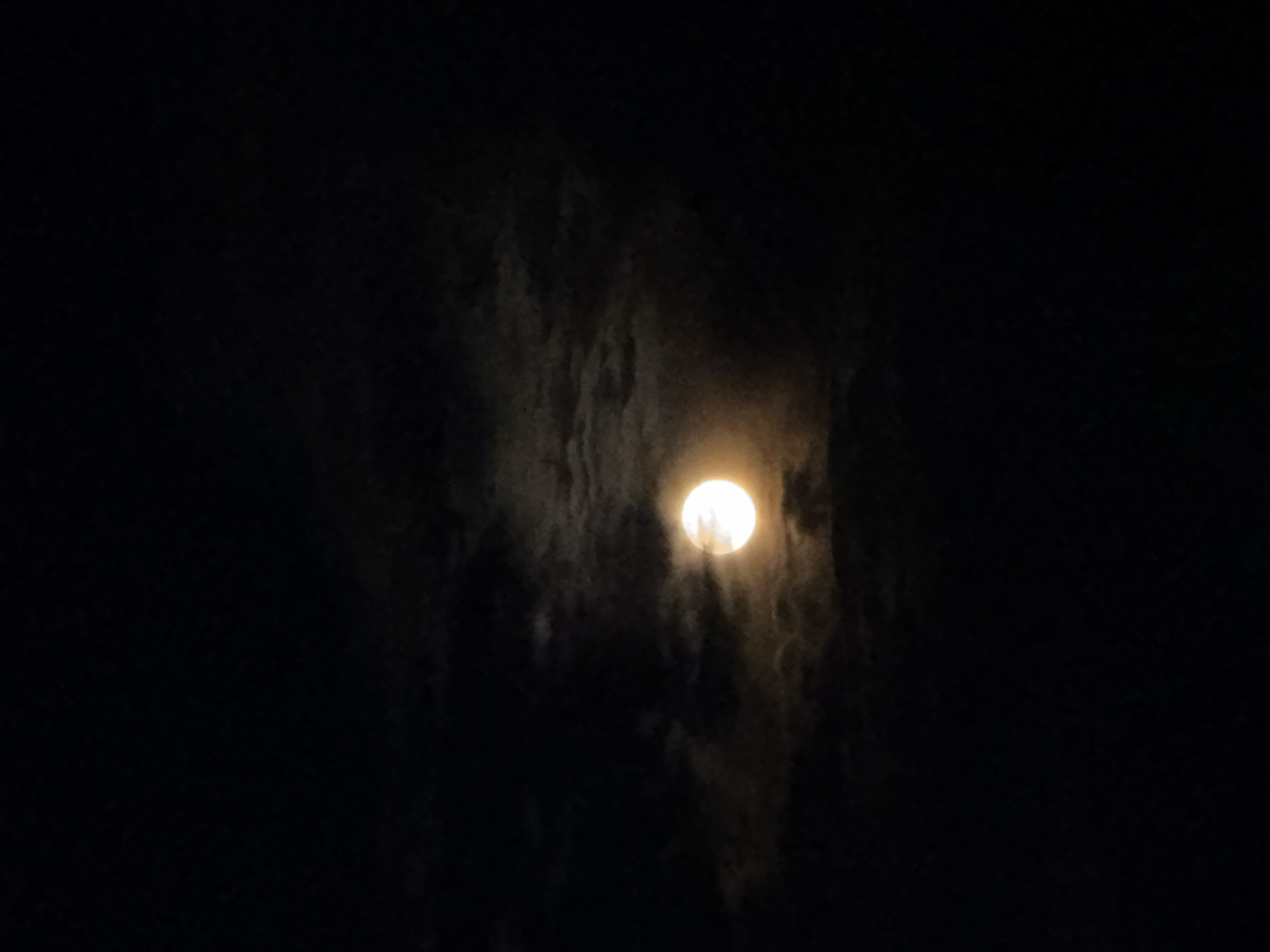 The image depicts a full moon, partially obscured by clouds. The setting is nighttime, with the surroundings appearing dark. The moon emits a bright glow that illuminates the surrounding clouds. The overall atmosphere is dark and somewhat mysterious. The location in the city of Chaikovsky, Russia is not discernible from the image. No people, subjects, objects, actions, or interactions are visible other than the moon and clouds.