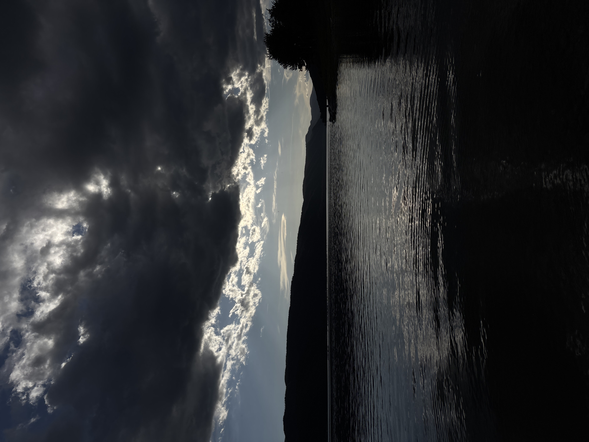 The image depicts a body of water bisected by a dark, silhouetted mountain range under a dramatic sky. To the left, a large mass of dark, heavy clouds dominates the view, partially obscuring patches of bright sky. The water reflects the varying light levels, ranging from subtly illuminated ripples to areas of deep shadow. A silhouette of trees is visible at the edge of the water. The location is Unknown, Unknown.