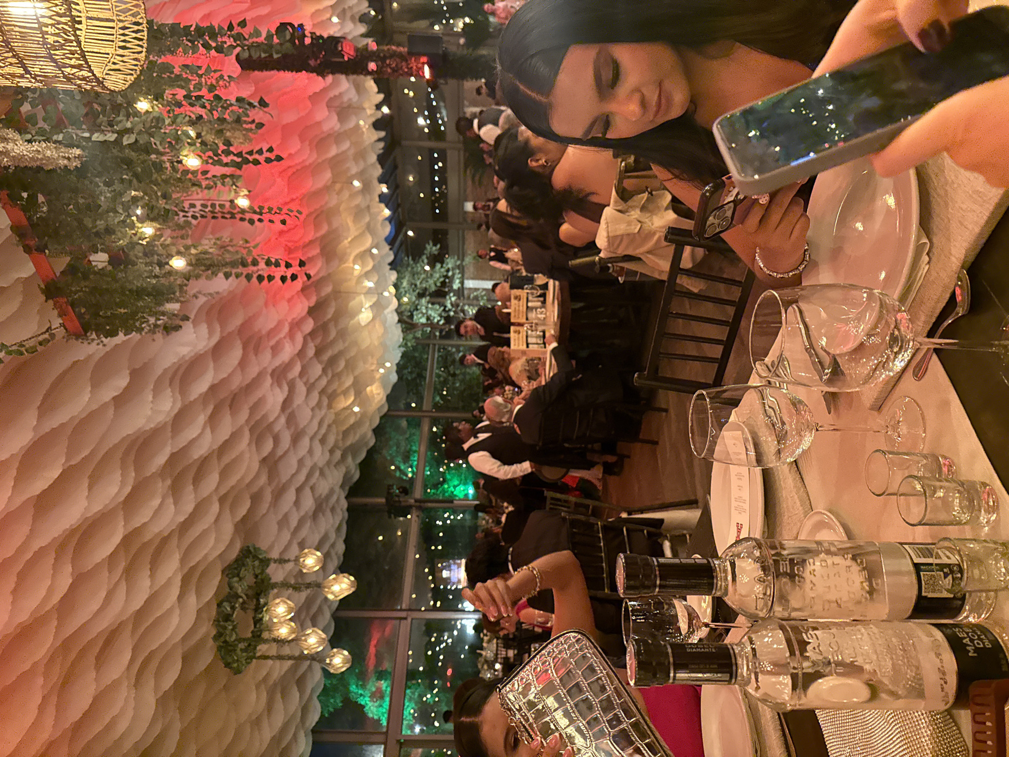 The image depicts a formal indoor dining event, likely a banquet or reception, held at "Salón Diamante" in San Andrés Cholula, Mexico. The ceiling is adorned with a decorative, cloud-like fabric structure, illuminated by hanging globe lights and lush green foliage. Red and green ambient lighting highlights the space.
In the foreground, a table is set with white plates, silverware, wine glasses, and two bottles of what appears to be tequila. Two small shot glasses are also present. To the left, a woman wearing a pink dress is visible, holding a silver, textured clutch bag. To the right, a young woman with dark hair is looking down at her phone, wearing a dark, strapless top and a diamond bracelet. Her hand holds a smartphone, and another hand, possibly belonging to another diner, is also visible holding a phone.
The background shows numerous guests seated at tables, some in formal attire. Servers in black vests are visible assisting guests. Large windows reveal an outdoor area with trees and decorative lights, suggesting the event is taking place at dusk or night. The overall atmosphere is one of celebration and elegance.