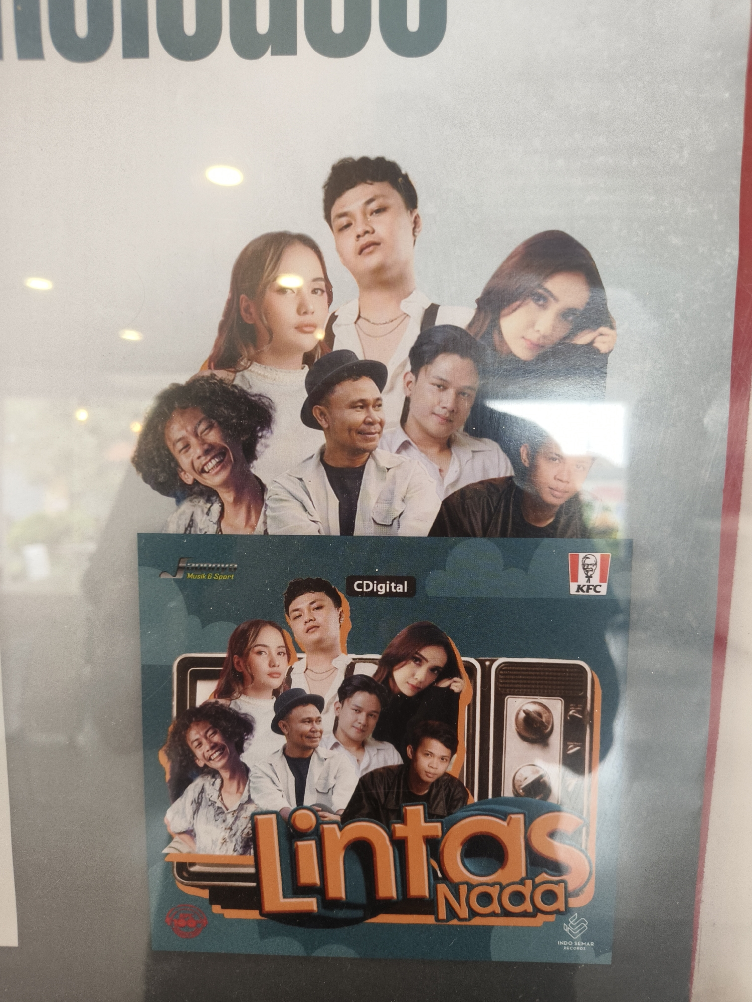 The image is a photograph of a poster or album cover featuring a group of people. At the top, there are several individuals arranged in a layered composition. In the lower portion of the image, a teal-colored cover with text and images is displayed. The text on the cover appears to be "LINTAS NADA" in large orange letters, with "Nada" in smaller text below. A KFC logo is visible on the right side of the cover. The lighting in the image suggests an indoor setting, possibly a store or an exhibition space, with reflections from overhead lights visible on the surface of the poster. There is no discernible information in the image that would suggest a specific location in Denpasar, Indonesia.