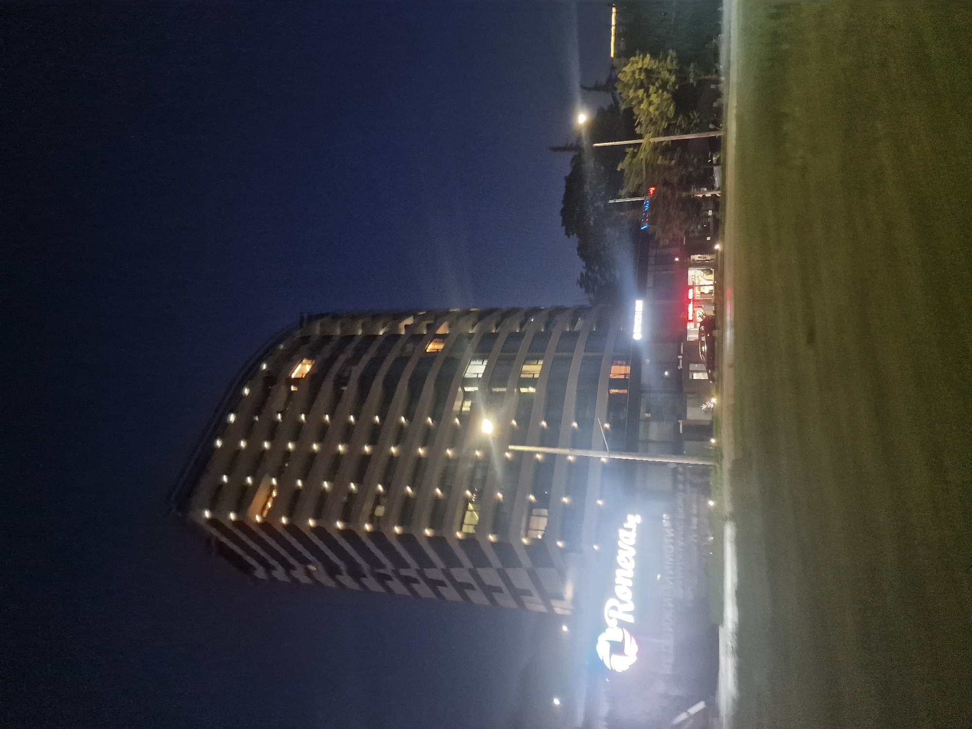 This is a nighttime photograph taken from an outdoor perspective, capturing a tall, modern, curved building in the city of Unknown, Unknown. The building is illuminated by numerous small, rectangular lights on each floor, arranged in a grid pattern. The structure has a dark, possibly black or dark grey, facade with lighter horizontal bands. The sky is a deep, dark blue, indicating nighttime.
In the foreground, a well-lit sign with the logo "Roneva" is visible, accompanied by smaller text beneath it. To the right of the building, a street lamp with a bright light shines upwards. Further to the right, trees are visible, illuminated by the ambient light and possibly the street lamp. Across the street from the building, there are other structures, including what appears to be a building with illuminated red signage and windows, suggesting commercial activity. A car is parked in front of this building. The ground in the foreground is covered in grass, which appears somewhat dimly lit. There is a slight atmospheric haze or fog contributing to the diffused lighting. No people are clearly visible in the scene.
