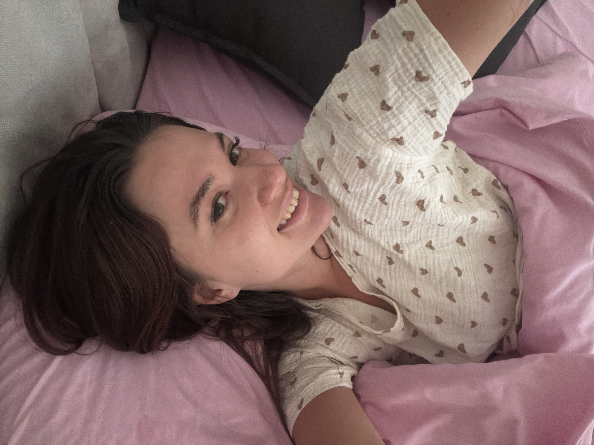 A single individual, a young adult female with medium-brown hair, is depicted lying down on a bed in an indoor setting. She is positioned with her head slightly tilted to the left, looking directly at the camera with a broad smile, exposing her upper and lower teeth. Her eyes are dark. She is wearing a light-colored, possibly off-white or cream, short-sleeved top adorned with numerous small, brown, heart-shaped patterns; the fabric exhibits a textured, crinkled appearance. She is partially covered by a plain pink duvet or blanket, which also serves as the pillow supporting her head. In the upper left background, a light grey, textured fabric surface is visible, likely part of a headboard or additional bedding. To the upper right, a dark grey or black surface is present. The scene is consistent with a domestic bedroom environment. The specific location within the city of Belgrade, Serbia, cannot be determined from the visual information provided in this image.