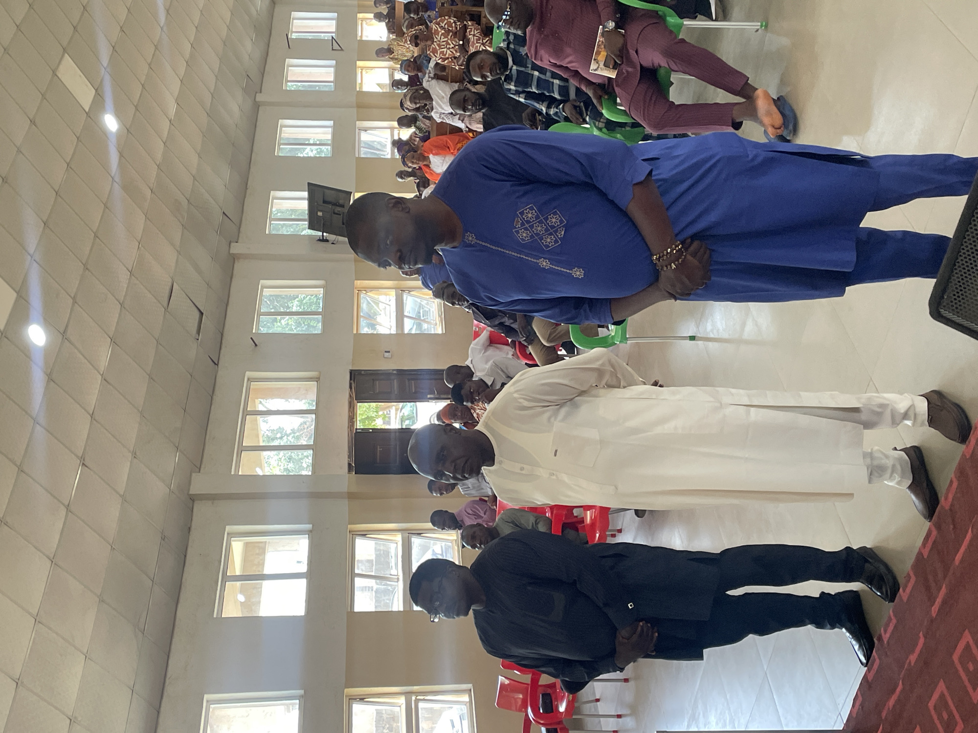 A gathering of people is observed indoors in Bukuru, Nigeria. Three men stand prominently in the foreground, each wearing different colors: blue, white, and black. Behind them, a larger crowd is seated. The architecture features multiple windows, providing natural light. The setting appears to be a large room, potentially a meeting hall.