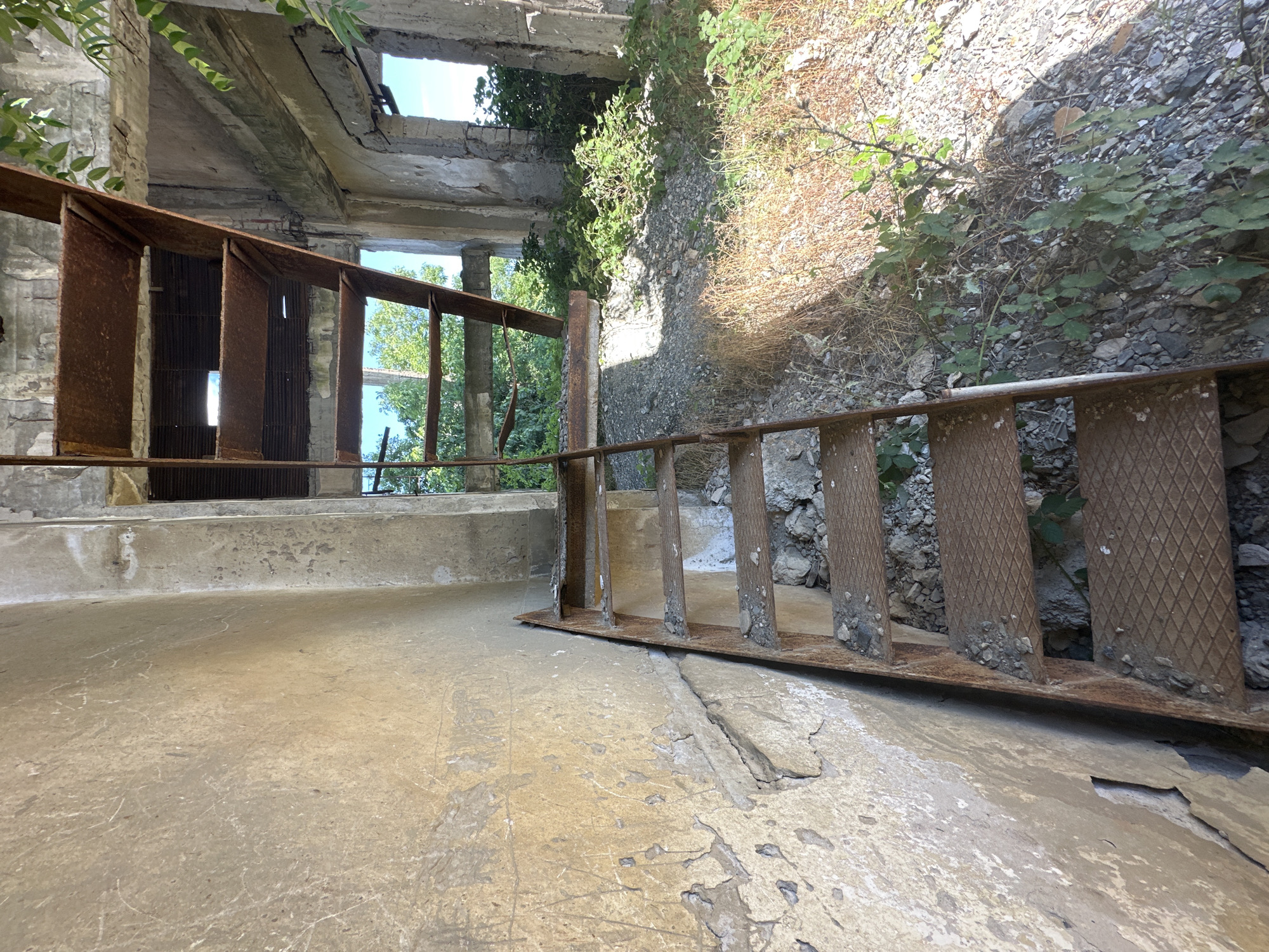 The image depicts an abandoned, partially collapsed concrete structure with a rusty metal staircase leading upwards. The walls of the structure are made of weathered concrete, with some sections showing peeling paint and graffiti. The ground is covered in rubble, dirt, and overgrown vegetation. Sunlight streams through openings in the roof and walls, illuminating the scene. The overall impression is one of decay and neglect. This is a view of an abandoned structure, possibly a former industrial building, located in or near the city of Rosen, Bulgaria. There are no people visible in the image.