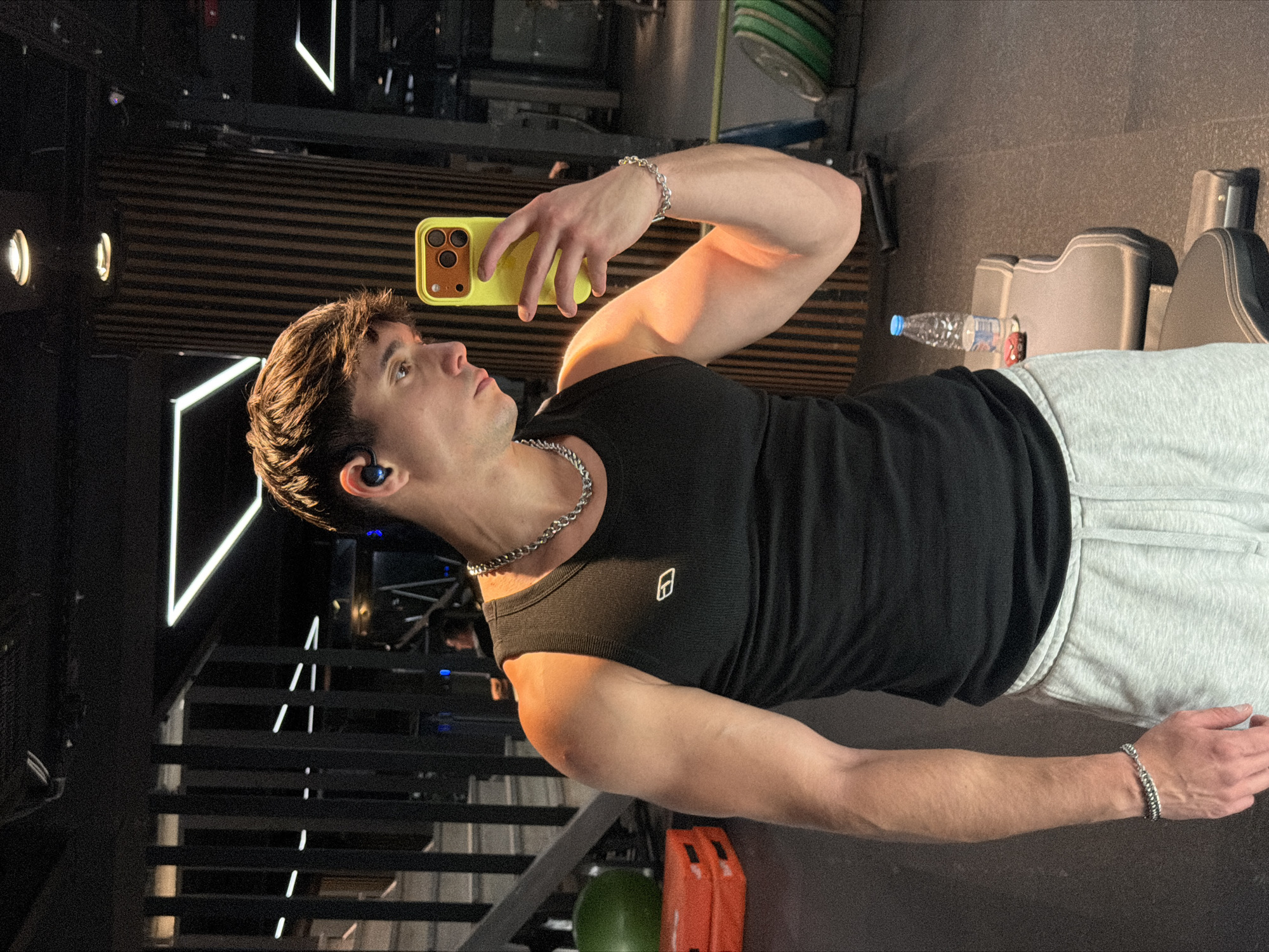 A young man is taking a selfie in what appears to be a gym. He is wearing a black tank top, grey sweatpants, a silver necklace and a bracelet. He is holding a yellow-cased iPhone up to take the photo. The environment includes weightlifting equipment, a water bottle, and modern interior design with linear lighting. The location is potentially in Sofia, Bulgaria, but no definitive landmarks or elements confirm it.