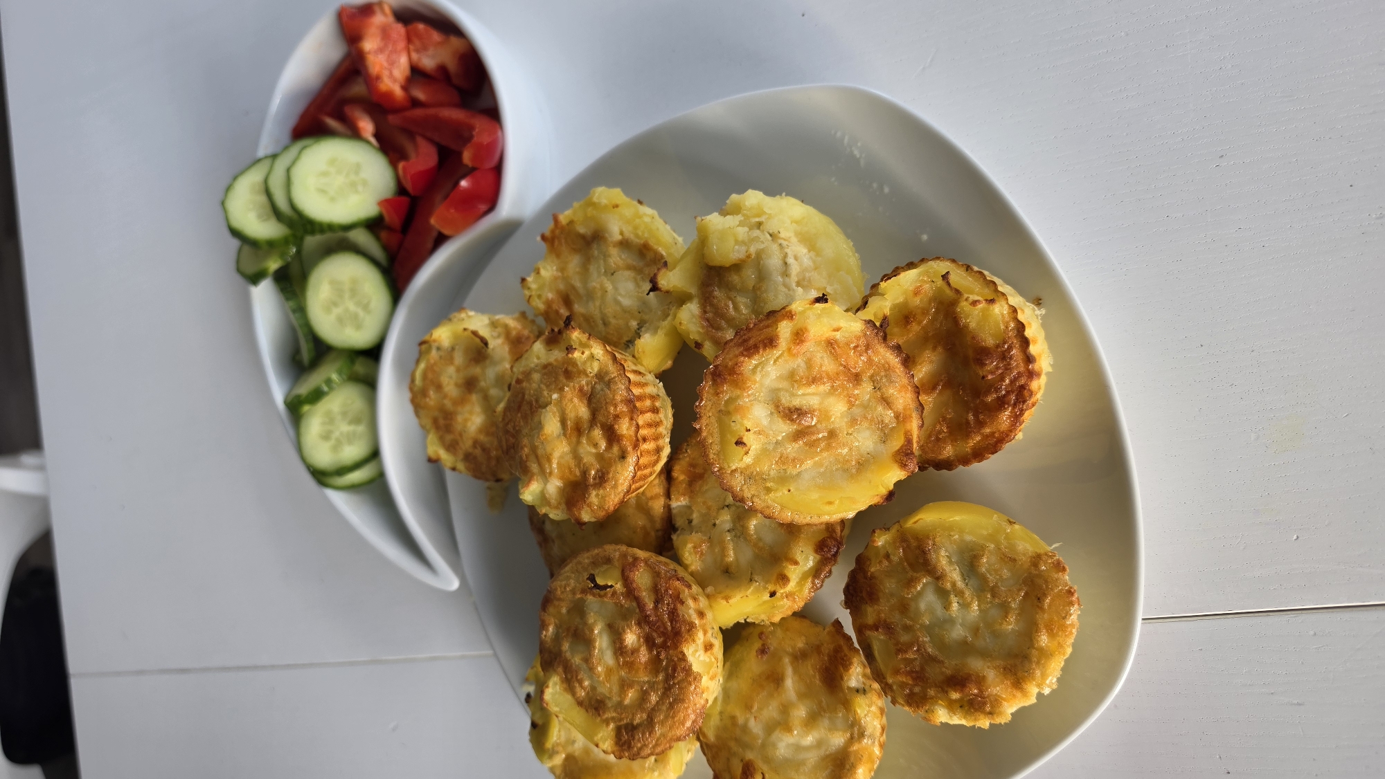 A top-down shot showcases a meal consisting of savory potato muffins or fritters served alongside a fresh vegetable salad. The main dish features approximately a dozen golden-brown, round potato items, some with ridged edges, arranged on a white, oval-shaped plate positioned on the right side of the frame. Their texture appears crispy on the outside.
To the left, a separate white, segmented serving dish holds the accompanying salad. One section of this dish is filled with neatly sliced green cucumbers, while the adjacent section contains chopped red bell peppers.
The entire arrangement rests on a clean, white tabletop, suggesting an indoor setting like a kitchen or dining area. The bright lighting, possibly from a window or overhead light, illuminates the food and surface, creating subtle shadows and highlighting the fresh appearance of the ingredients. No people or text are visible in the image.