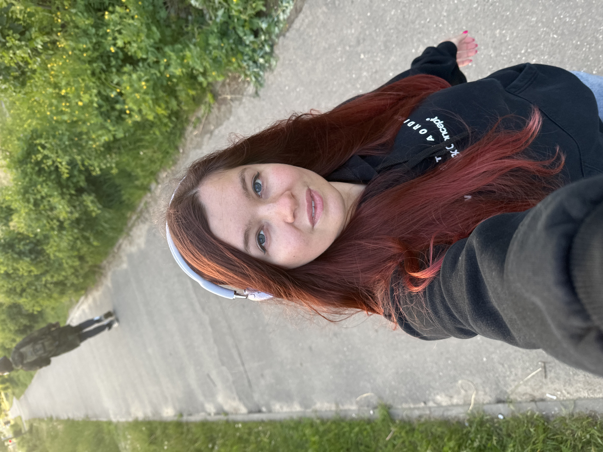 A person with auburn hair and blue eyes takes a selfie outdoors. They are wearing a black hoodie and white headphones. The person is standing on a concrete path with green vegetation on either side. In the distance, a person's shadow can be seen on the path. The location is Vladimir, Russia.