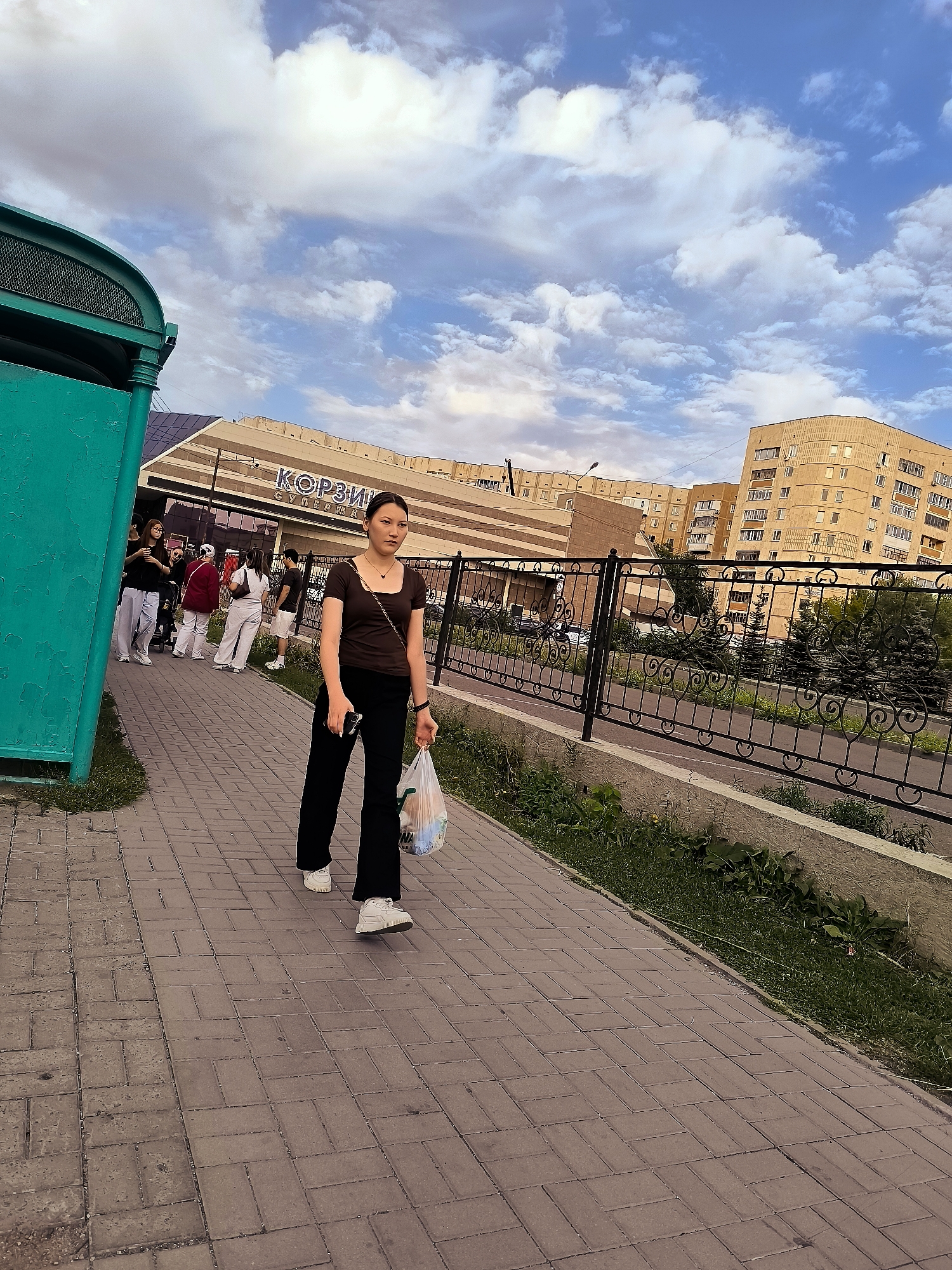 A young woman walks down a brick-paved sidewalk in Karaganda, Kazakhstan, carrying a clear plastic bag with some items in it and a phone in her hand. She is wearing a dark brown short-sleeved shirt and black pants. The sidewalk is bordered by grass and a decorative metal fence. In the background, there is a large building with a sign that reads "КОРЗИ" (KORZI) in large letters, a supermarket, and several apartment buildings. The sky is cloudy with patches of blue. Several other people are visible further down the sidewalk, some standing and some walking. A teal colored structure is on the left side of the image.