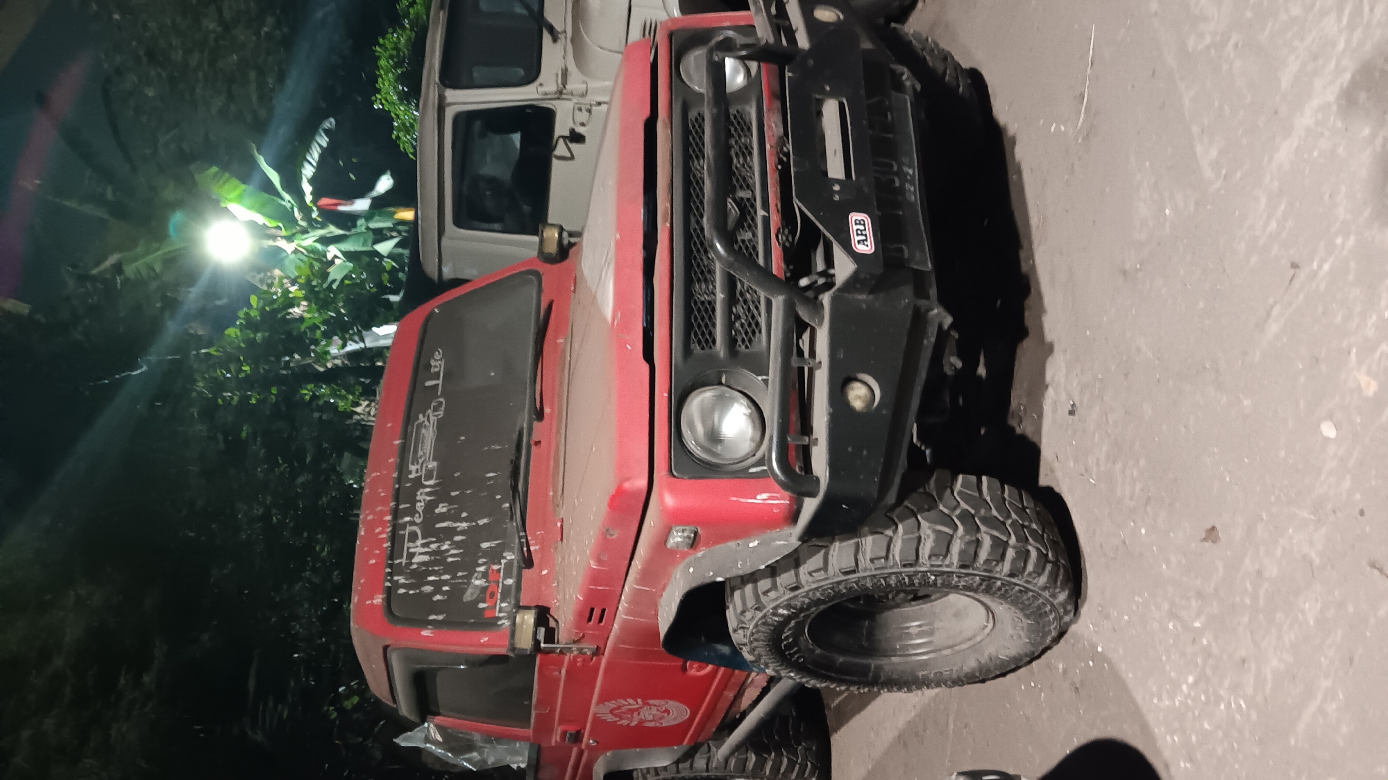 The image shows a red, four-wheel-drive vehicle, possibly a Suzuki Samurai, parked outdoors at night in what appears to be a yard or parking lot. The vehicle is dusty and has some white streaks on the windshield. It is equipped with off-road tires and a bull bar on the front. Another vehicle, lighter in color and with a boxy shape, is partially visible behind it. The background consists of trees and foliage, with a bright light source illuminating the scene from above, casting strong shadows. The ground is paved and appears to be asphalt or concrete. There are no people visible in the image.