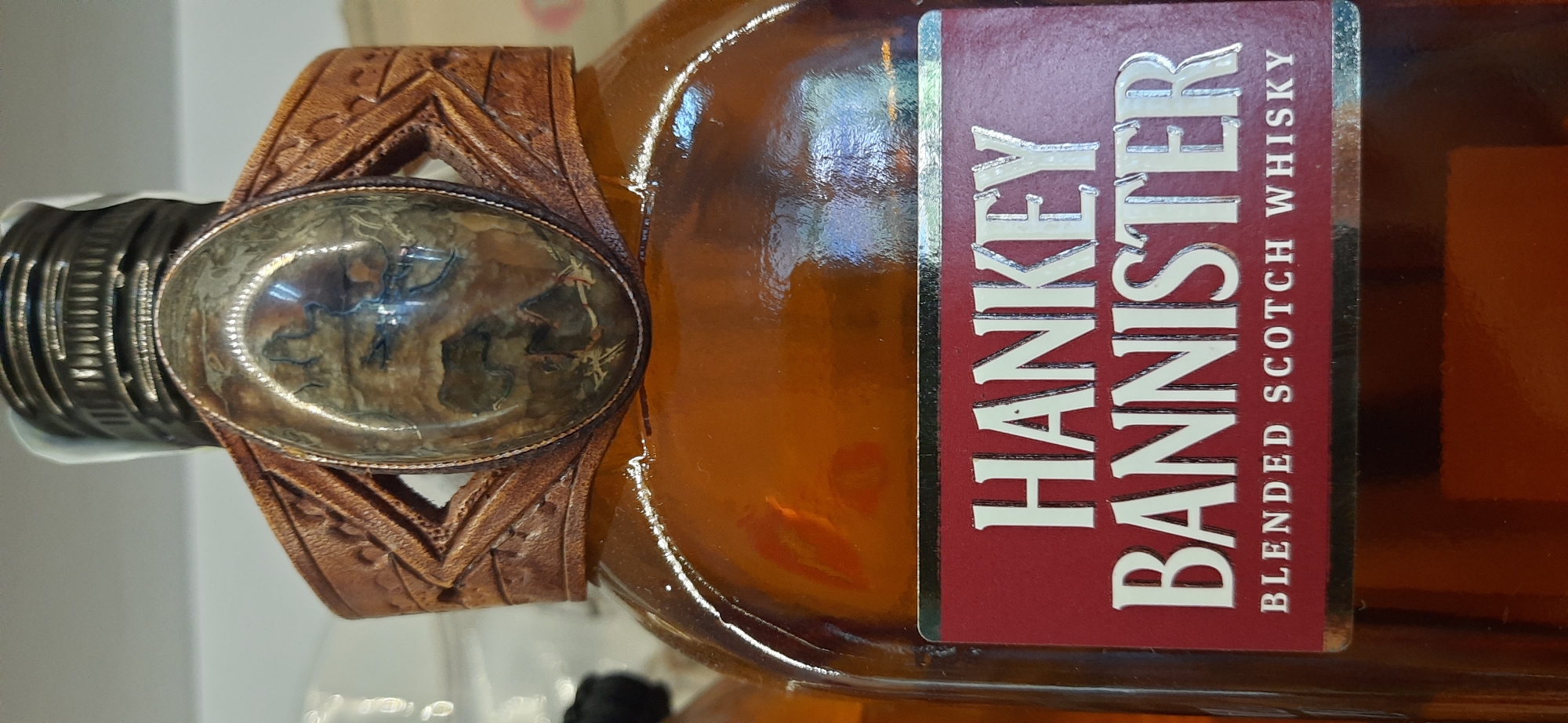 A close-up, eye-level, medium shot captures a decorative amber glass bottle of Hankey Bannister blended Scotch whisky. The bottle's neck is adorned with a leather cuff featuring intricate geometric embossing and a large, oval, multi-colored polished stone, possibly agate or jasper, set in a metallic bezel. Below the cuff, a red label with silver metallic lettering reads "HANKEY BANNISTER BLENDED SCOTCH WHISKY." The background is blurred and appears to be a retail environment with shelves and other products. The lighting is bright, creating reflections on the glass bottle. The location in Dnipro, Ukraine, is not discernible from the visual information provided.