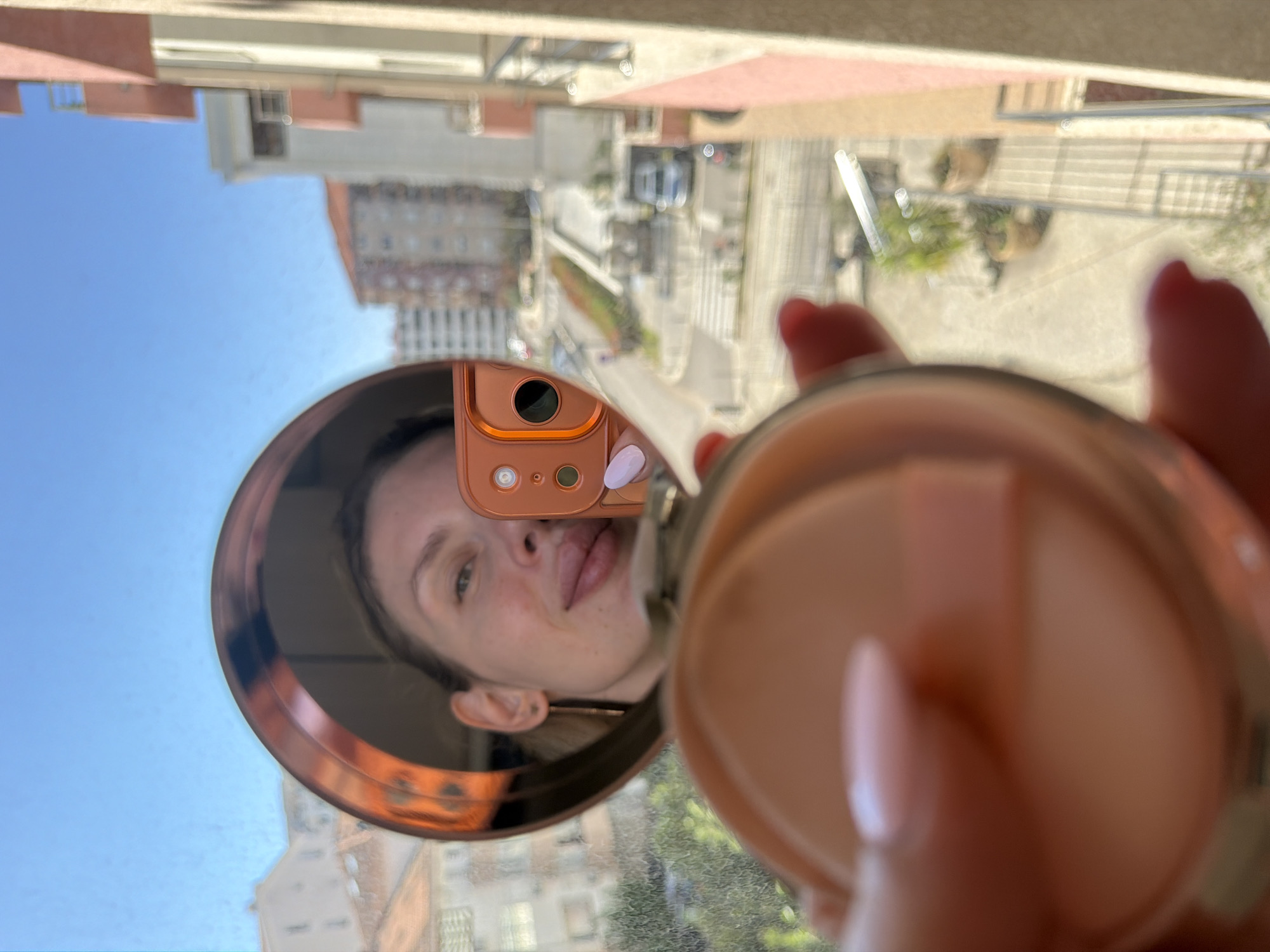 The image, rotated 90 degrees counter-clockwise, captures a mirror selfie taken from an elevated position, likely a balcony or window, overlooking an urban landscape in Budva, Montenegro.
In the foreground, a hand with light pink manicured fingernails holds an open, peach-toned makeup compact, with a soft white puff visible inside, slightly blurred. The central subject is a woman's face, reflected in a round mirror with a copper-colored frame. She has her tongue slightly extended in a playful or casual expression. An orange-cased smartphone, used to capture the image, is also visible within the mirror's reflection, held by her hand.
Behind the mirror and visible through the window/balcony is a clear, bright blue sky dominating the upper portion of the background. Below the sky, an urban scene unfolds with multi-story buildings featuring typical Mediterranean architecture, including terracotta-colored elements. A paved street with faint markings and some sparse greenery can be seen in the distance, suggesting a sunny daytime. There is no visible text in the image.