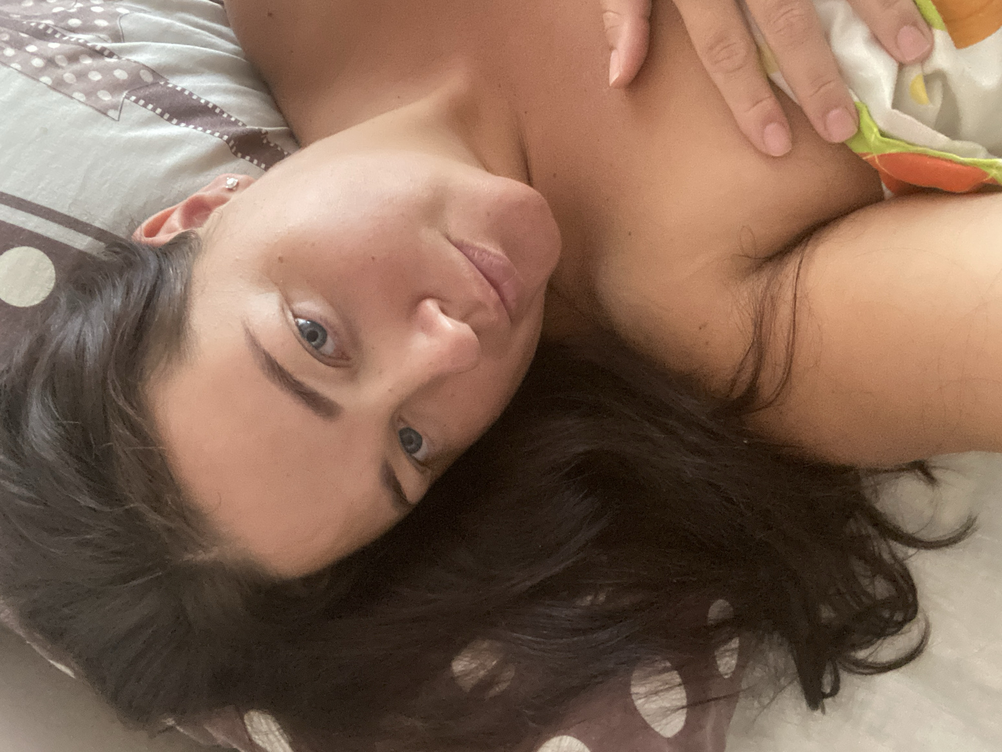 A woman with dark hair and blue eyes is lying in bed, looking at the camera. She is wearing a small diamond earring in her left ear. Her right hand is resting on her chest, and she is partially covered by a colorful blanket. The background is a pillow with a patterned design. The image appears to have been taken indoors. There is no information in the image to suggest the location is Sochi, Russia.