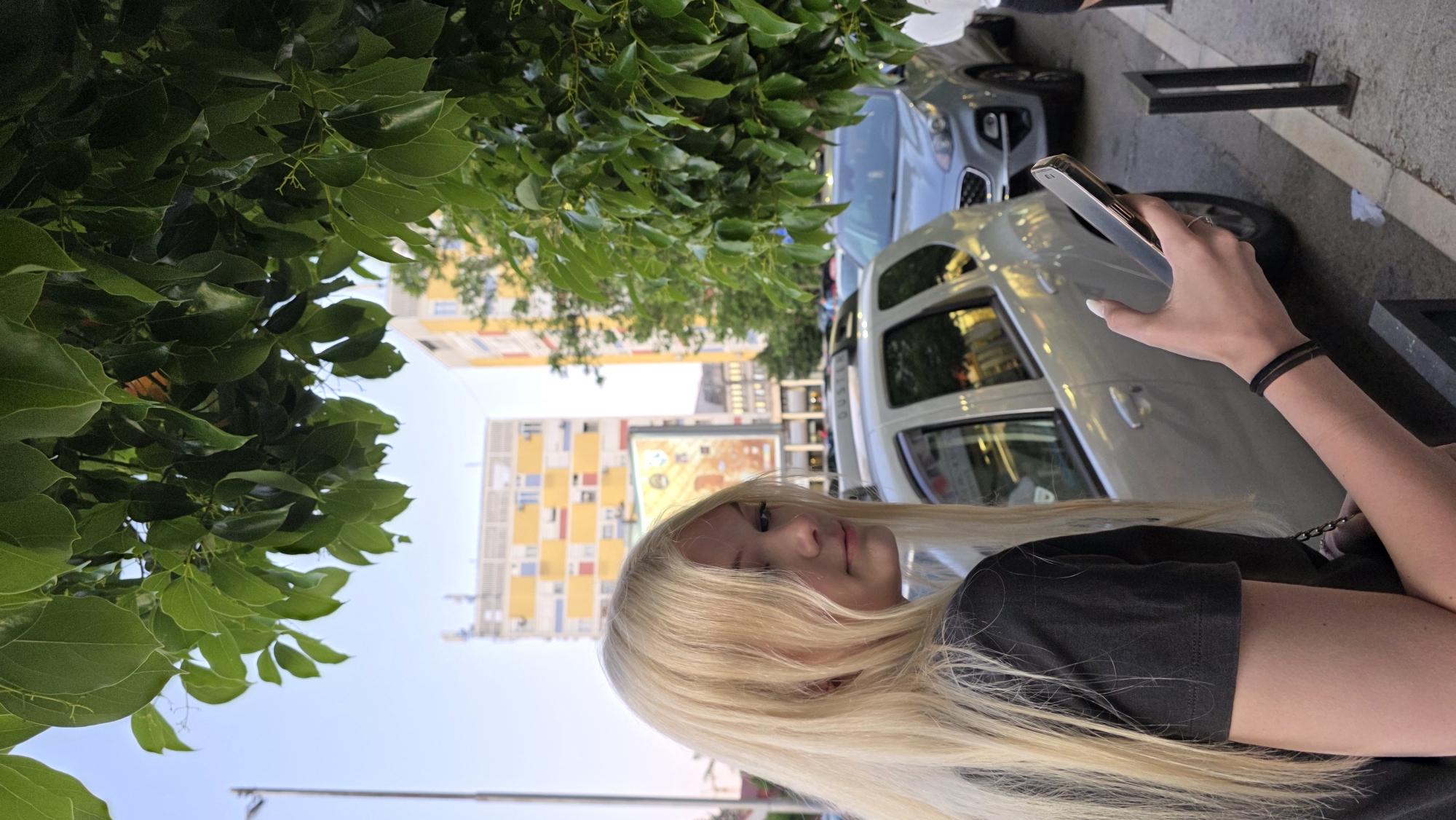 A young woman with long blonde hair is looking at the camera. She is wearing a black t-shirt. She is holding a silver smartphone in her right hand. In the background, there are several parked cars, a tree with green leaves, and an apartment building. The apartment building is white with yellow, orange, and blue square accents. The location appears to be in Podgorica, Montenegro.