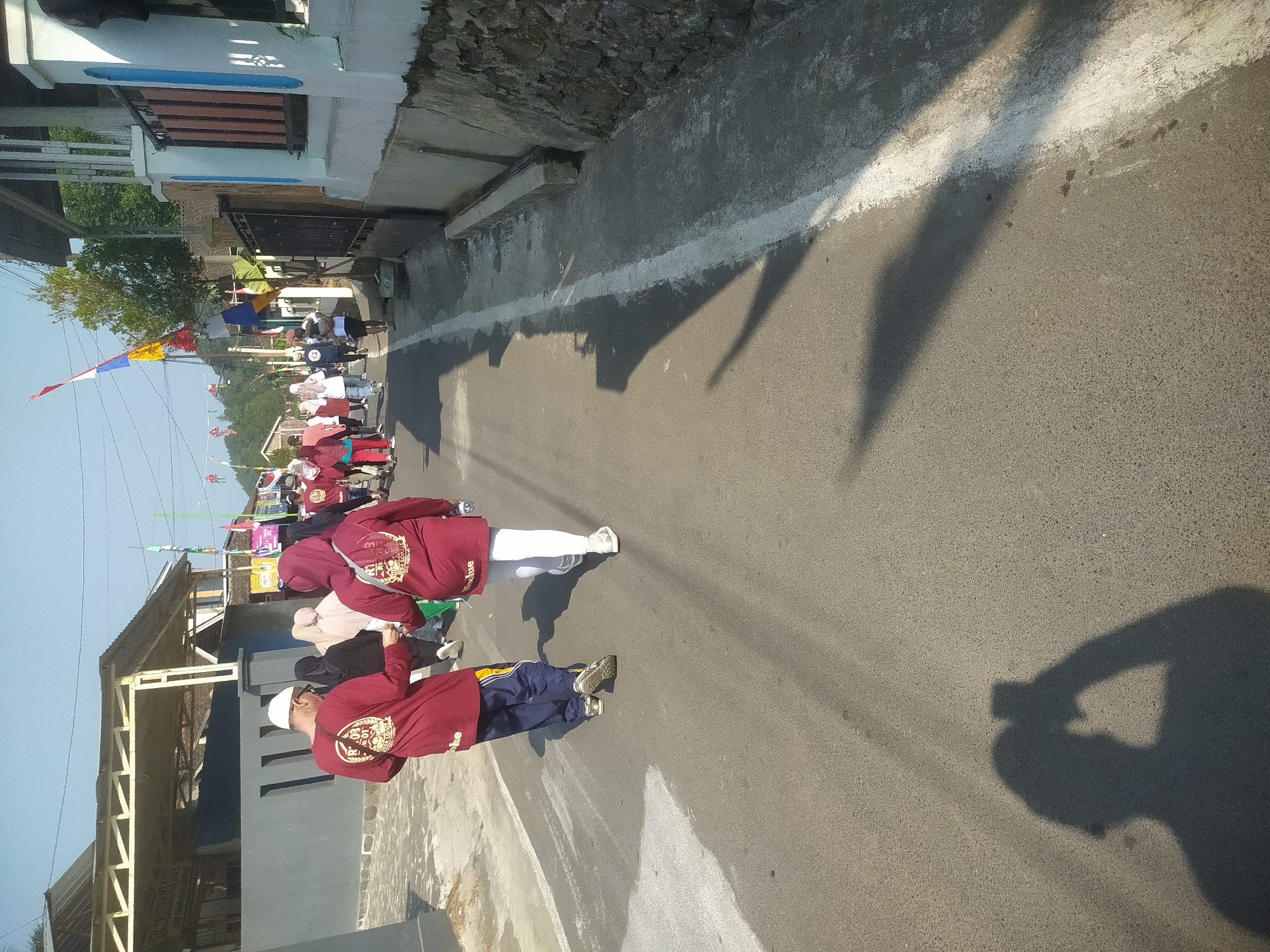 A group of people are walking down a paved road in Lopait, Indonesia, under a clear blue sky. The people are wearing predominantly maroon colored clothing, some with white headscarves, suggesting a communal event or activity. In the foreground, two individuals are walking away from the camera, one man wearing a white cap and a maroon long-sleeved shirt with an emblem on the back, and a woman in a maroon hooded sweatshirt and white pants. The road is lined with buildings and vegetation on either side. Flags and banners are visible in the background, adding a festive or celebratory atmosphere. The sun casts long shadows on the road, indicating it is either early morning or late afternoon. The environment appears to be a residential area with a mix of traditional and modern architectural elements.
