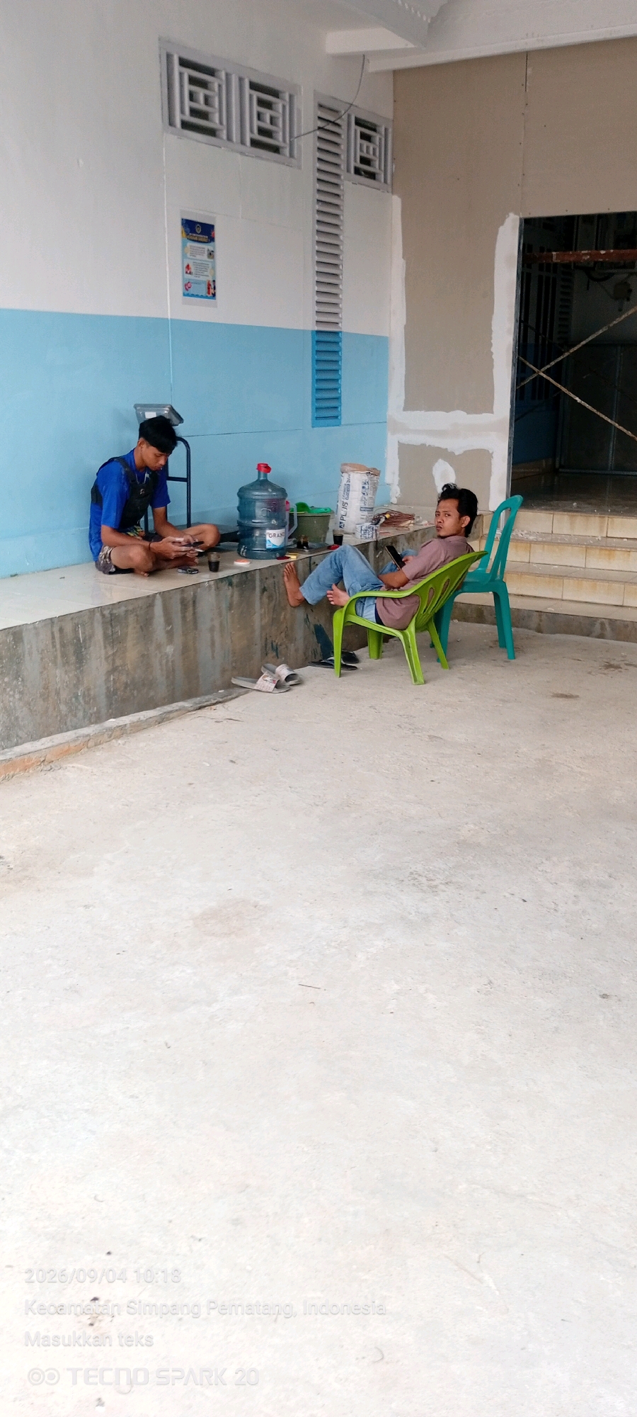 In Simpang Pematang, Indonesia, two young men are present in an outdoor, possibly under-construction or renovation area. One man sits cross-legged on a raised concrete ledge, engaged with his phone. He wears a blue vest over a dark shirt and shorts. Beside him on the ledge are a large water cooler, a bag of what appears to be cement, and some cups. The other man is seated in a lime-green plastic chair, also looking at his phone, with his legs crossed. He wears a purple shirt and blue jeans. A teal plastic chair is positioned behind him. The environment features a concrete floor and walls painted white and light blue, with metal grilles and louvers integrated into the upper portion of the wall. Stairs are visible in the background to the right. A date stamp and location information, "2026/09/04 10:18, Kecamatan Simpang Pematang, Indonesia," are overlaid on the image.