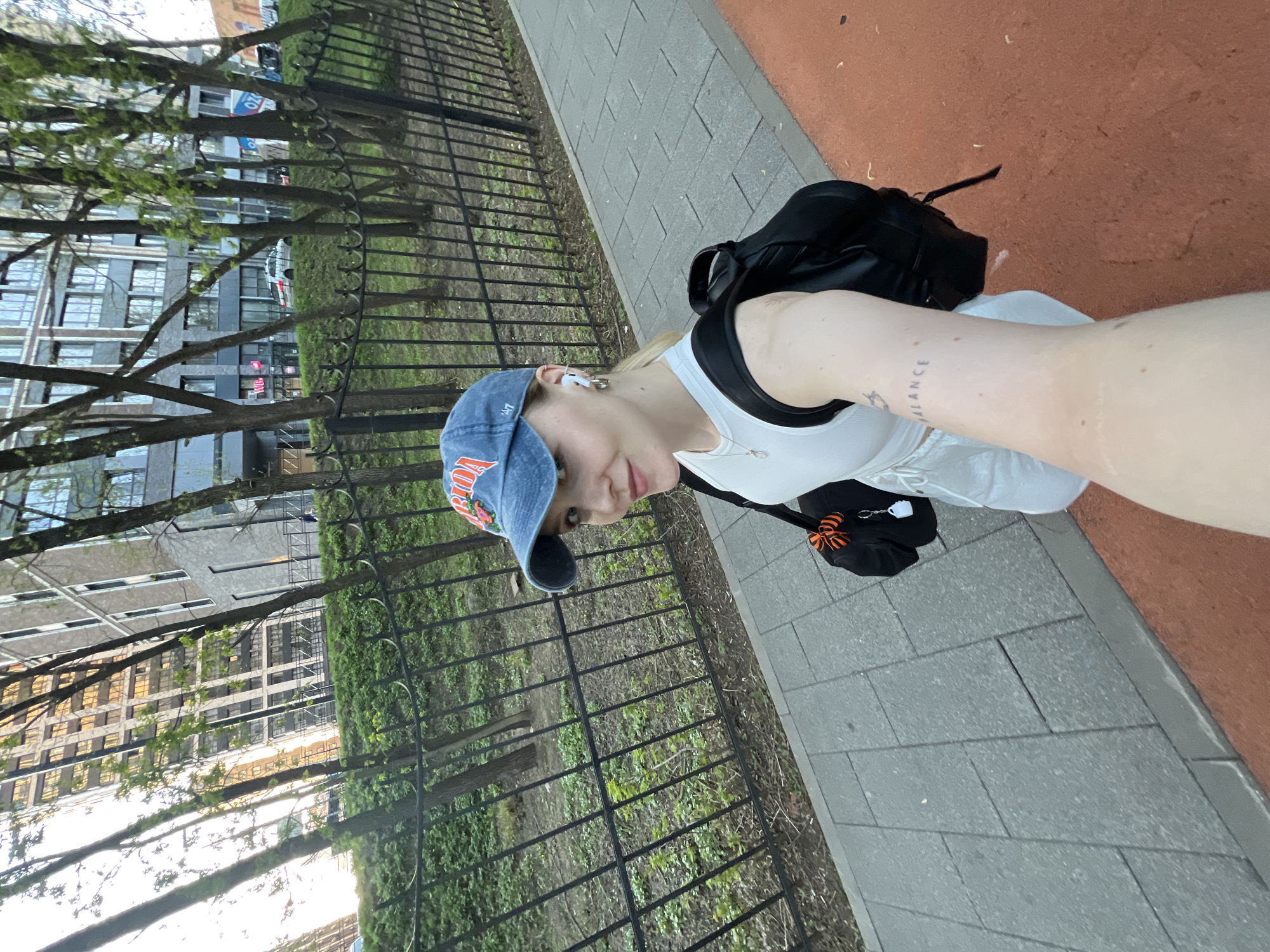 A woman is taking a selfie while walking on a sidewalk next to a metal fence and a grassy area with trees. She is wearing a blue baseball cap, a white top, and a black backpack. Her hair is blonde, and she is wearing white earbuds. A tattoo is visible on her inner forearm. The background includes buildings and trees. The sidewalk is made of gray concrete blocks, and there is a red, rubberized surface adjacent to it.