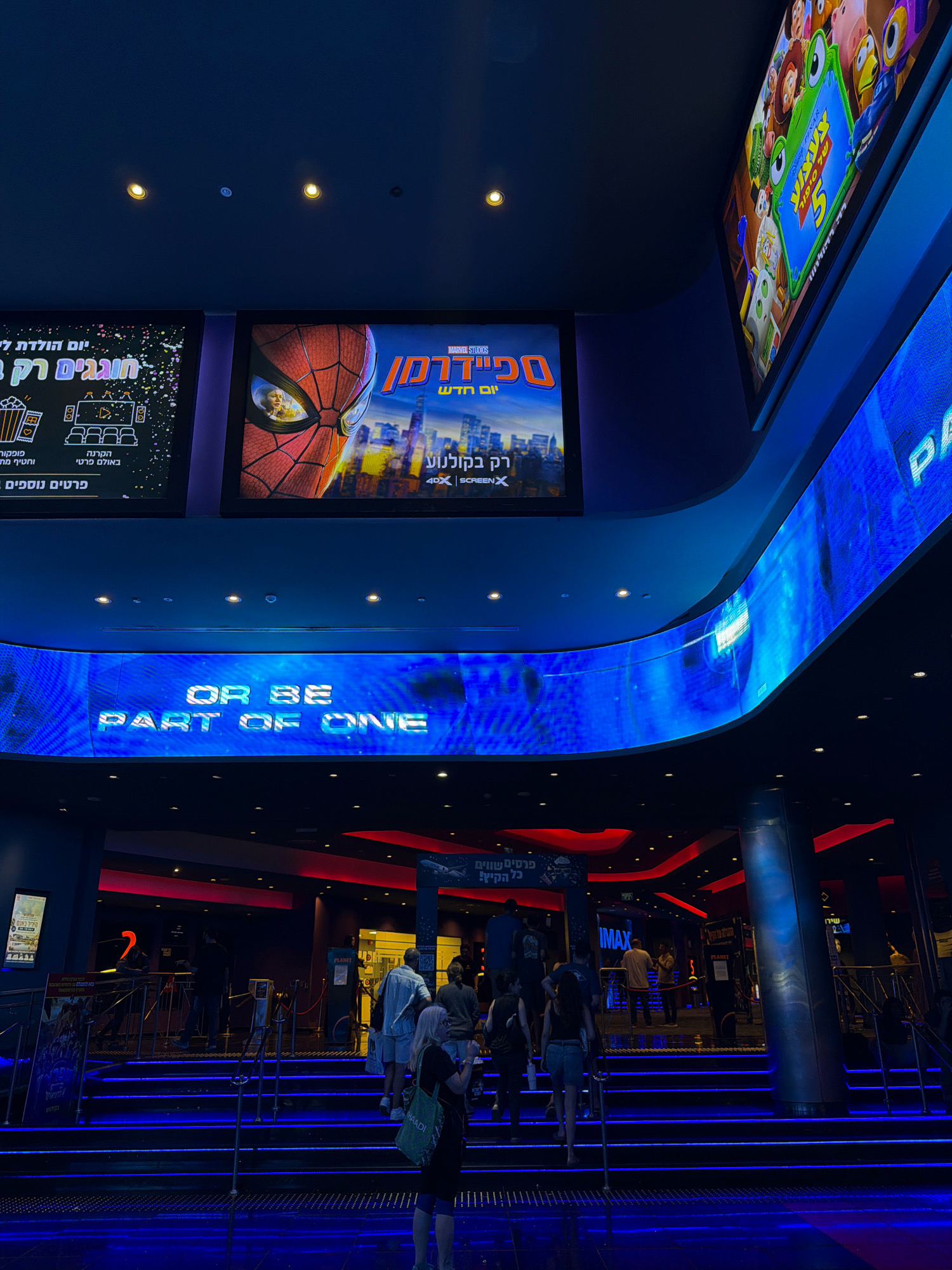 The image depicts the interior of a modern cinema, likely located in Rishon LeZion, Israel, given the Hebrew text visible on promotional displays. The scene is dominated by a futuristic ambiance, with extensive use of blue LED lighting illuminating the entrance and cascading down the steps.
In the upper portion of the frame, large digital screens showcase movie posters. One screen prominently features a close-up of Spider-Man's mask, with Hebrew text below indicating it's a Marvel Studios production. Another screen displays characters from the Disney Pixar movie "Toy Story 5" in Hebrew. A third, curved screen spans across the mid-ground, displaying the English phrase "OR BE PART OF ONE" in a futuristic font against a blue, textured background.
The lower section of the image shows the entrance to the cinema complex, with people ascending a wide, illuminated staircase. Several individuals are seen walking up the steps, some carrying bags. The entrance is flanked by railings and ticket booths. The overall lighting is dim, with the blue LEDs providing the primary illumination, creating a dramatic and immersive atmosphere. The architecture appears contemporary, with dark ceilings and metallic accents. Promotional posters and signage in Hebrew are visible at various points, suggesting the location's geographical context.