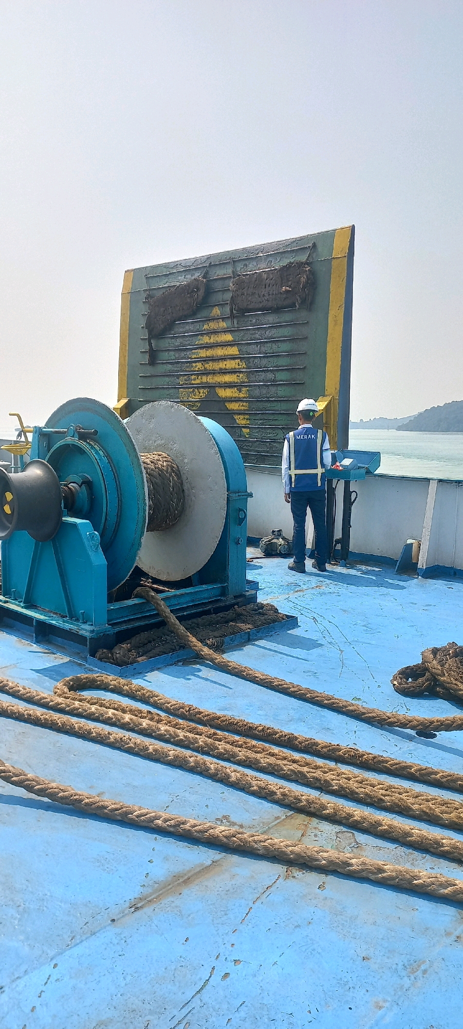 The image depicts a deck of a ferry in Bakauheni, Indonesia, under a clear sky. In the foreground, thick, weathered ropes lie coiled and stretched across the blue painted deck, indicating readiness for docking procedures. To the left, a large, teal-colored winch, likely used for handling mooring lines or anchors, dominates the scene. Its drum is partially wrapped with a heavy rope. In the mid-ground, a person wearing a white hard hat and a blue vest with "MERAK" printed on the back stands near a control panel. This individual appears to be a crew member or port worker. Behind them, a large, green and yellow metal structure, possibly part of the ferry's ramp or a docking guide, rises prominently. The background shows a body of water, with a hilly coastline visible in the distance, suggesting a maritime setting within or near Bakauheni.