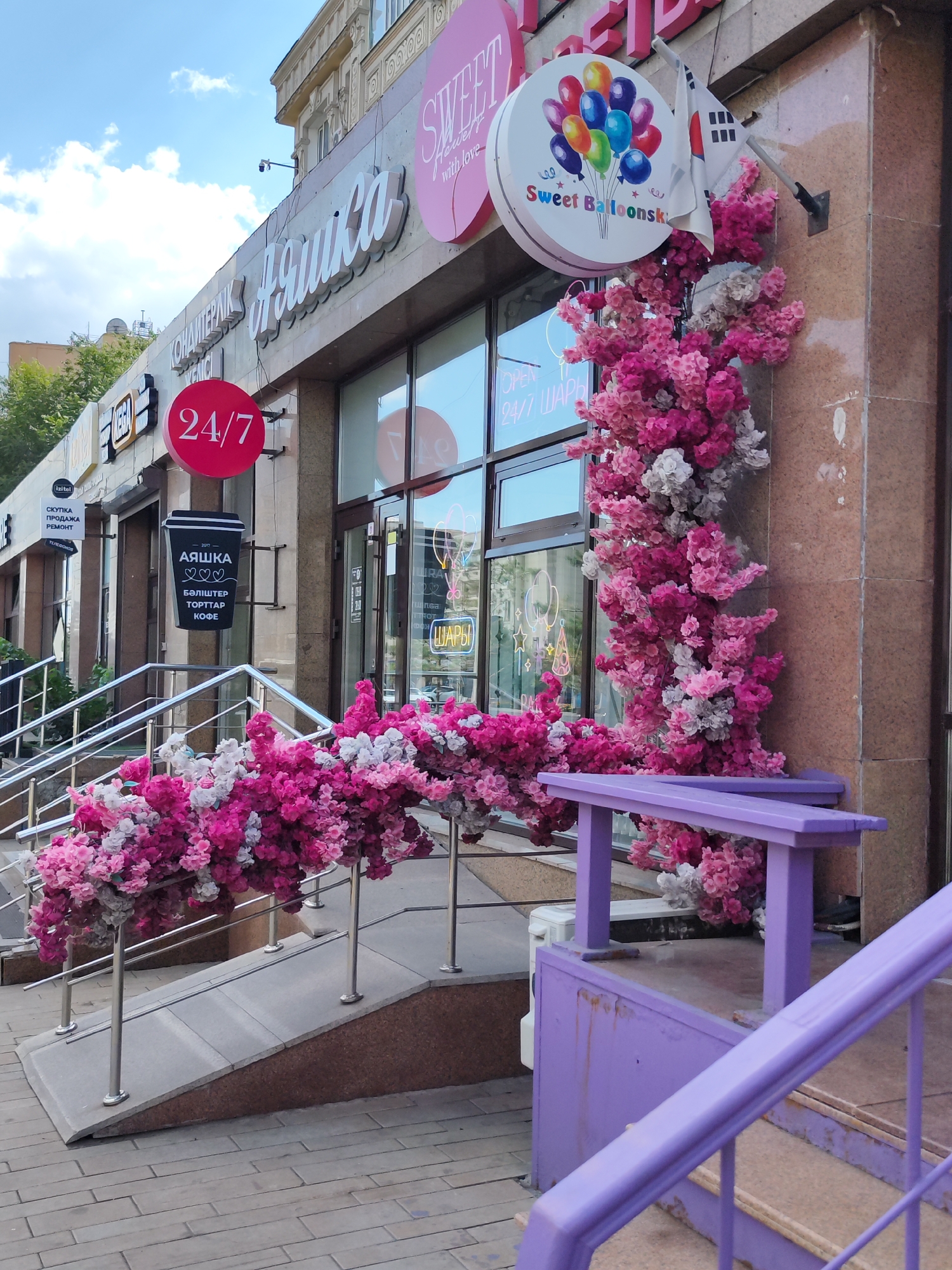 The image shows the exterior of a building with a bright pink floral decoration cascading down the facade and along a ramp. The building's signage includes "SWEET" in pink script, a circular sign with balloons and the text "Sweet Balloonsky," and another sign with Cyrillic text that appears to be for a confectionary or bakery, with the word "АЯШКА" (Ayashka) prominent. A red circular sign displays "24/7". The facade is made of textured brown stone, and large windows reflect the sky. A purple bench and railing are also visible, decorated with more of the pink and white floral arrangement. The area appears to be a commercial district, with cobblestone pavement in the foreground. The presence of a South Korean flag suggests a connection or affiliation. The scene is set in Astana, Kazakhstan.