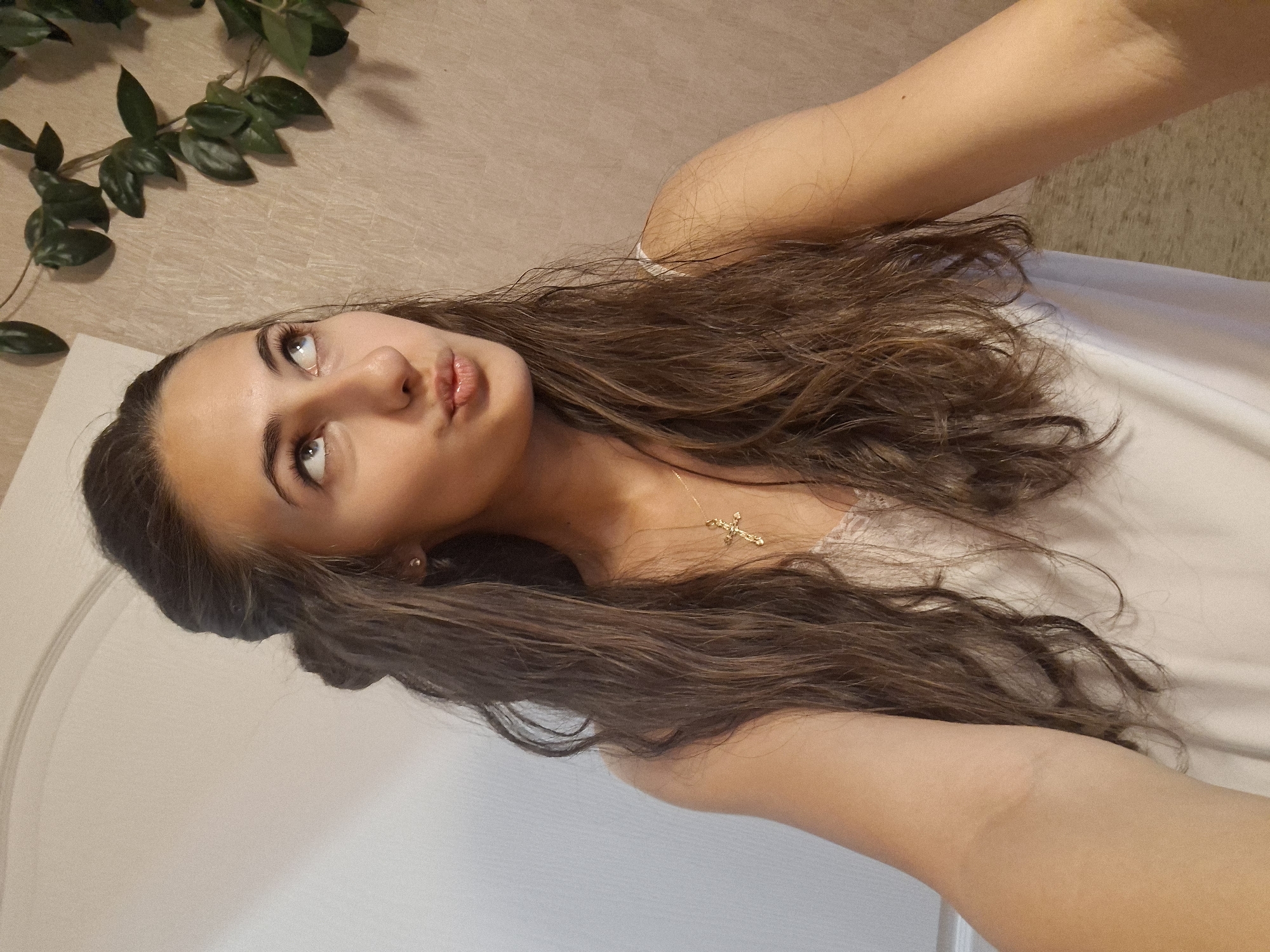 A young woman with long, wavy brown hair is pictured in a medium shot. She is wearing a white camisole with lace trim and a gold cross necklace. Her hair is partially pulled back, and she is looking upwards and to the right, with her lips puckered. She is positioned in front of a wall with a light, textured wallpaper, and a door is visible on the left side of the frame. A trailing plant with dark green leaves is on the upper right side of the image. The lighting appears to be artificial and coming from the front.