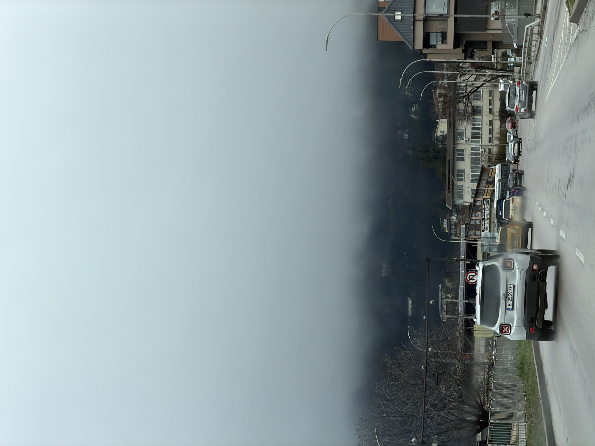 The image depicts a street scene in Varna, Bulgaria, partially obscured by dense fog or low clouds. A line of traffic is visible on a wet road, with various cars and a bus moving along the right side of the frame. A silver Jeep with the license plate B 2559 XE is prominently featured in the foreground. Buildings of varying heights, including apartment complexes and commercial structures, are situated along the road, some appearing partially shrouded in fog. Streetlights line the road, their visibility diminished by the weather conditions. The environment suggests a damp, overcast day with limited visibility due to the fog.