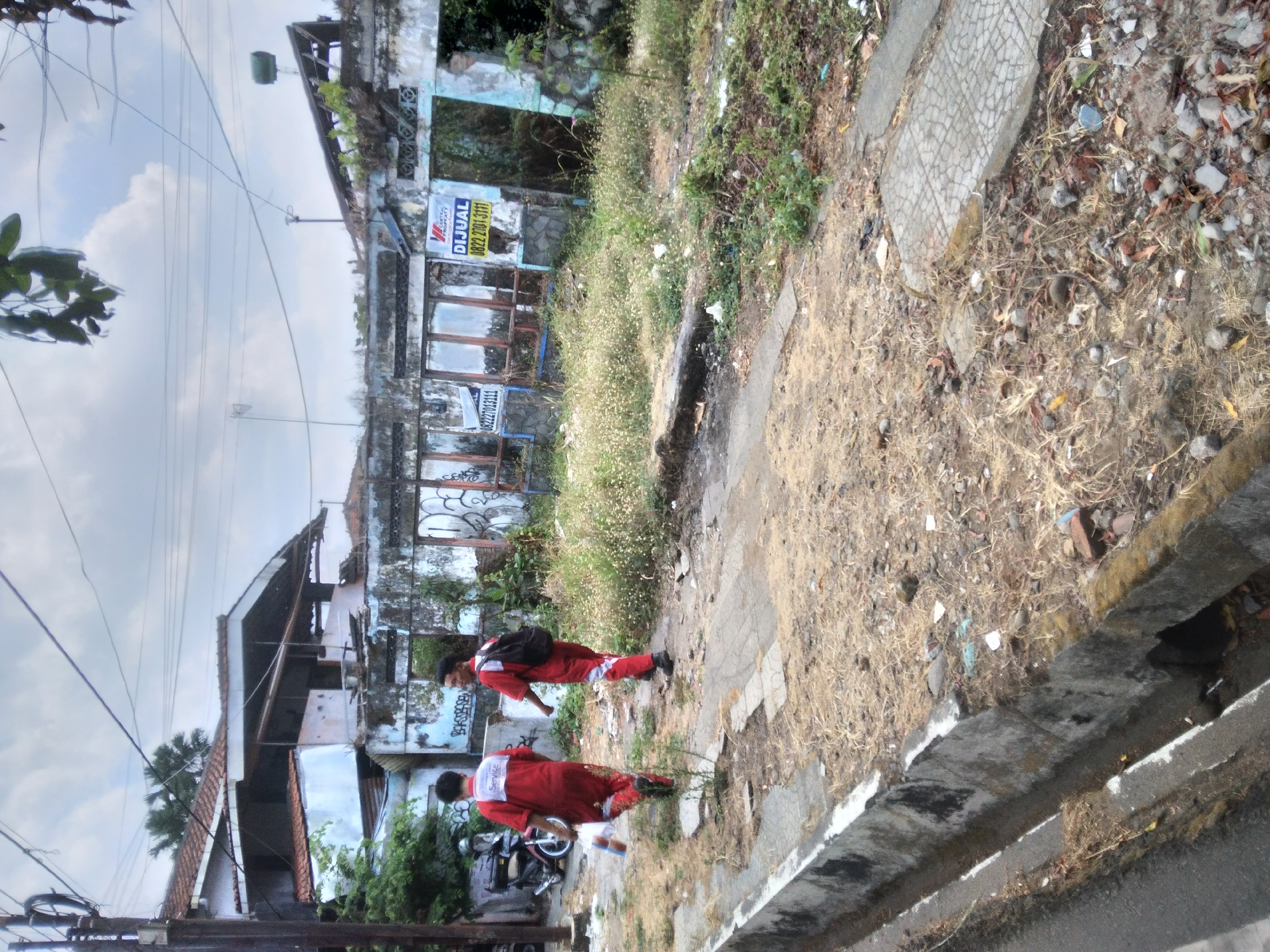 Two individuals wearing red jumpsuits are walking away from the camera along a dirt path. The person on the left is carrying plastic bags in their right hand and has "Service" printed on the back of their jumpsuit. The person on the right has a black backpack. They are walking past a row of dilapidated buildings, some with broken windows and graffiti. A motorcycle is parked to the left of the frame. The environment appears neglected, with dry grass, scattered debris, and overgrown vegetation. The scene is set outdoors, under a cloudy sky, with visible power lines overhead. The location is identified as Sokaraja, Indonesia.