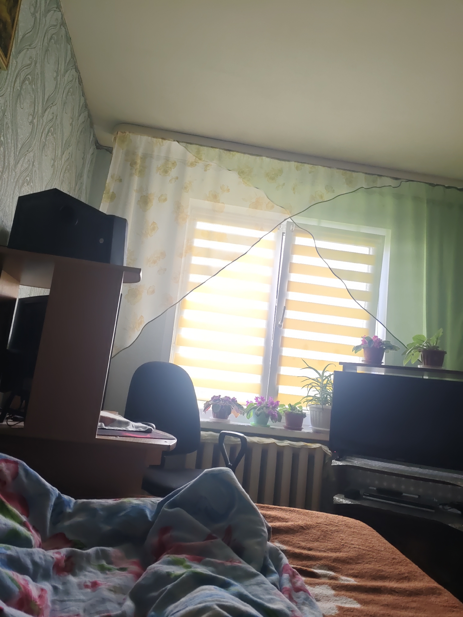 The image depicts an indoor scene, likely a bedroom or living area, within a residence in Kurashim, Russia. A window with striped blinds is prominent, letting in bright, yellow light. Several potted plants, including violets, are arranged on the windowsill. To the left of the window, a desk with a black speaker on top is visible, and a black office chair is positioned nearby. In the foreground, a person is partially covered by a floral patterned blanket and an orange blanket. To the right of the window, a television set is placed on a stand. The room's walls are adorned with patterned wallpaper and green curtains. A radiator is situated below the windowsill. A black wire runs diagonally across the upper portion of the image, connected to a light fixture. The overall impression is of a lived-in, domestic space.