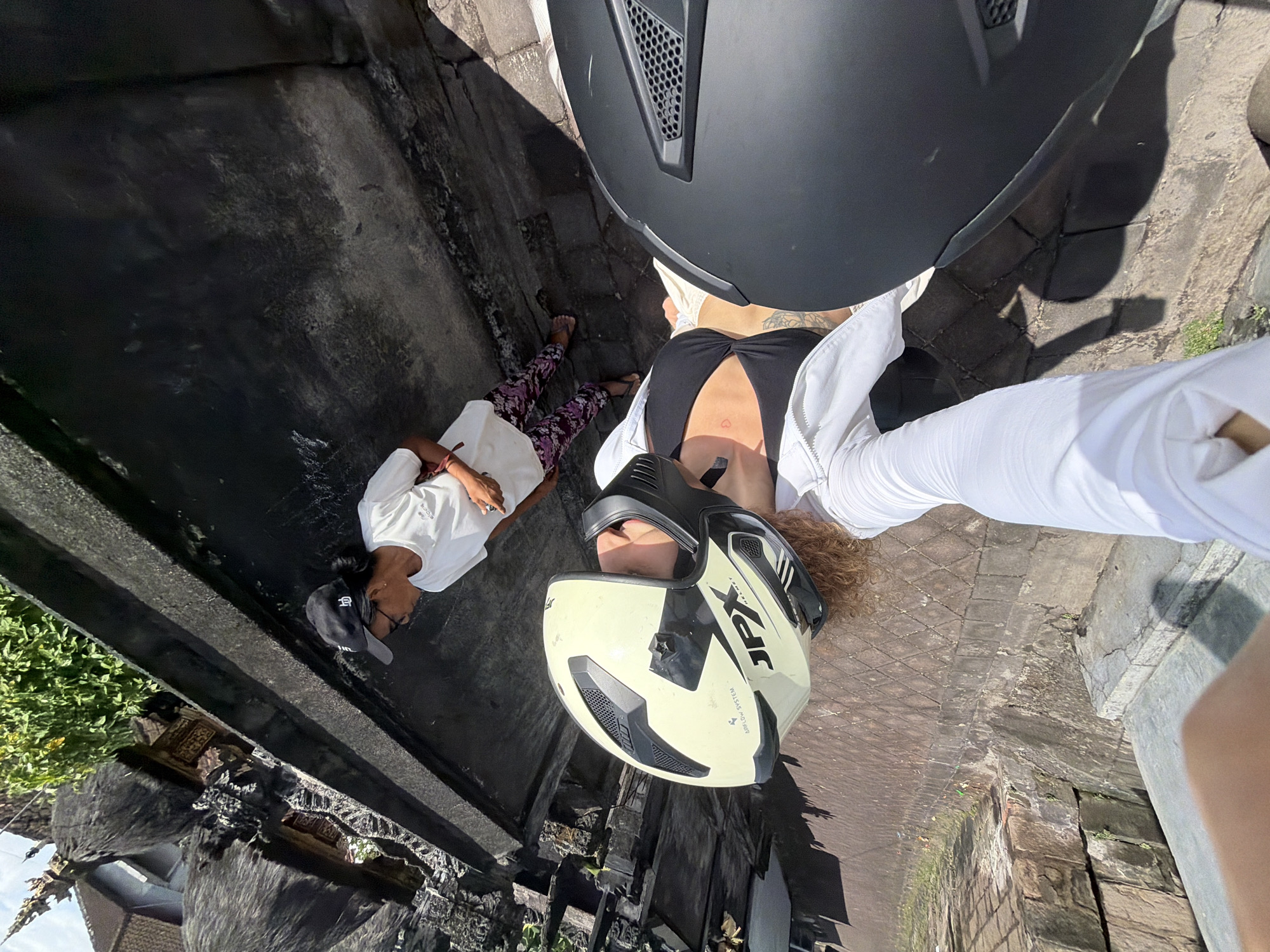 In Mengwi, Indonesia, a selfie captures two individuals on a stone pathway, bathed in bright sunlight. The person in the foreground is wearing a white jacket over a black swimsuit and a light yellow motorcycle helmet. Their left arm, clad in white, extends towards the camera. Their right hand, near their chest, has a small red mark. Beside them is a black motorcycle helmet. In the background, a person wearing a white t-shirt, patterned pants, and a black baseball cap stands looking down. The environment features dark, textured structures, possibly part of a temple or old building, with lush green foliage visible in the upper left corner. The ground is paved with stones.