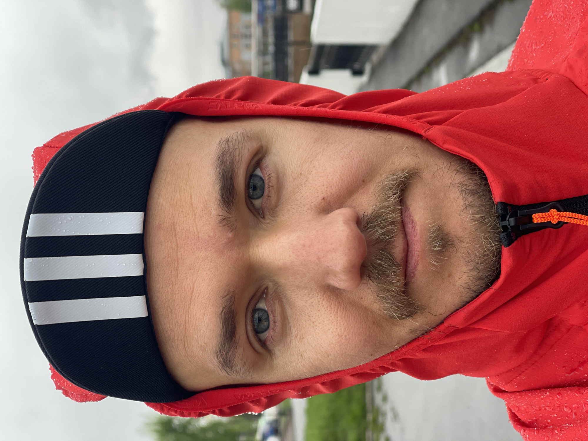 A man with a fair complexion, blue eyes, and light stubble is looking at the camera. He is wearing a black cycling cap with three white stripes and a red hooded jacket. The jacket is wet, with water droplets visible on the fabric. The background appears to be an outdoor urban setting, possibly in the city of Izhevsk, Russia.