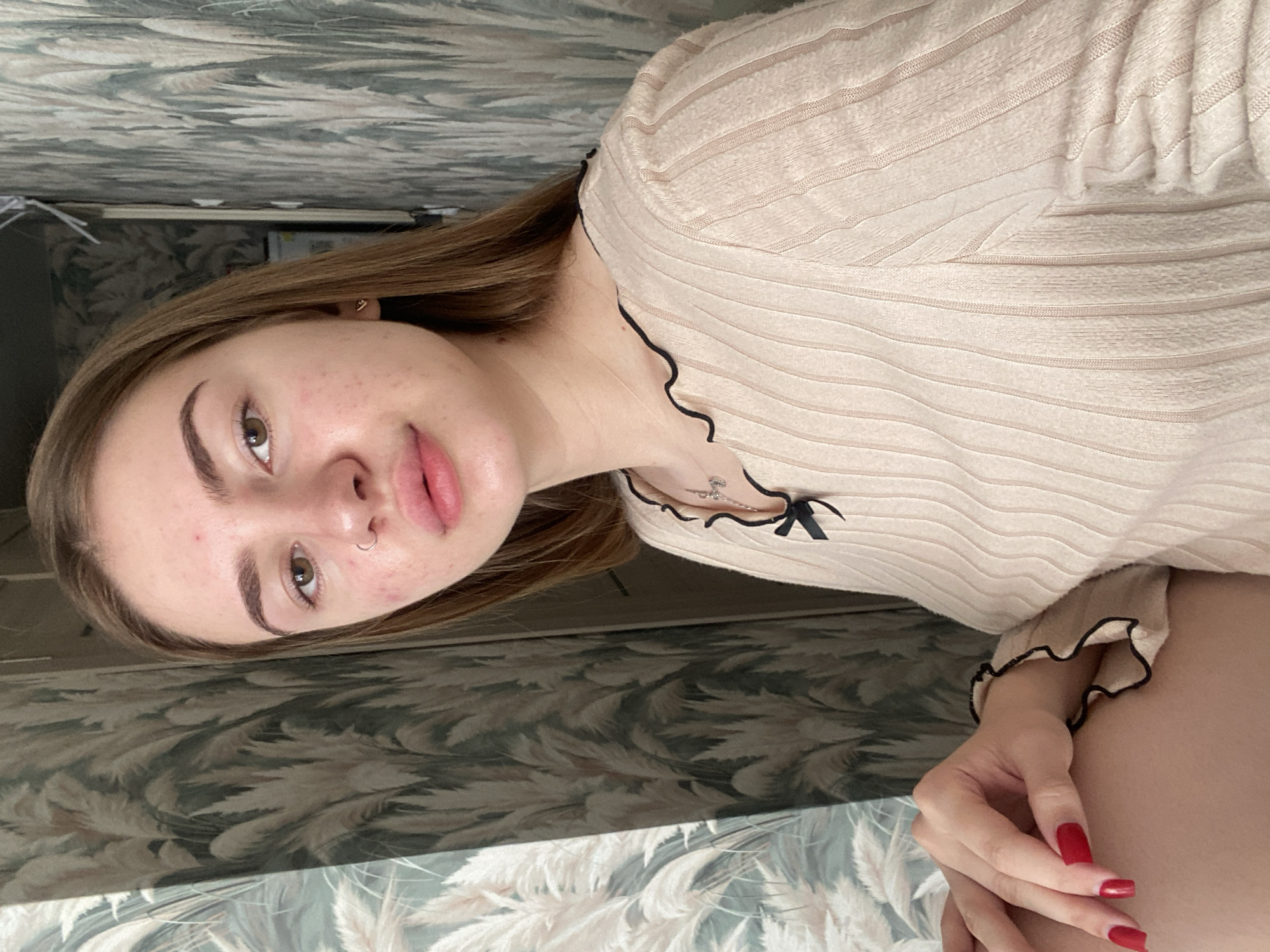 The image shows a close-up, waist-up, indoor portrait of a young woman with light brown hair, who appears to be in her late teens or early twenties. She is looking directly at the camera with a neutral expression. Her skin shows some redness and blemishes. She is wearing a light-colored, ribbed knit sweater with a black scalloped trim and a small black bow detail at the neckline. A delicate necklace with a small charm is visible around her neck. Her left hand is resting on her thigh, and her fingernails are painted bright red. The background consists of a patterned wallpaper with a natural motif, featuring tall, feathery plants in shades of green and beige. The lighting suggests an indoor setting, possibly a bedroom or living area. There is no indication of the location being in the city of Дьяково, Russia.