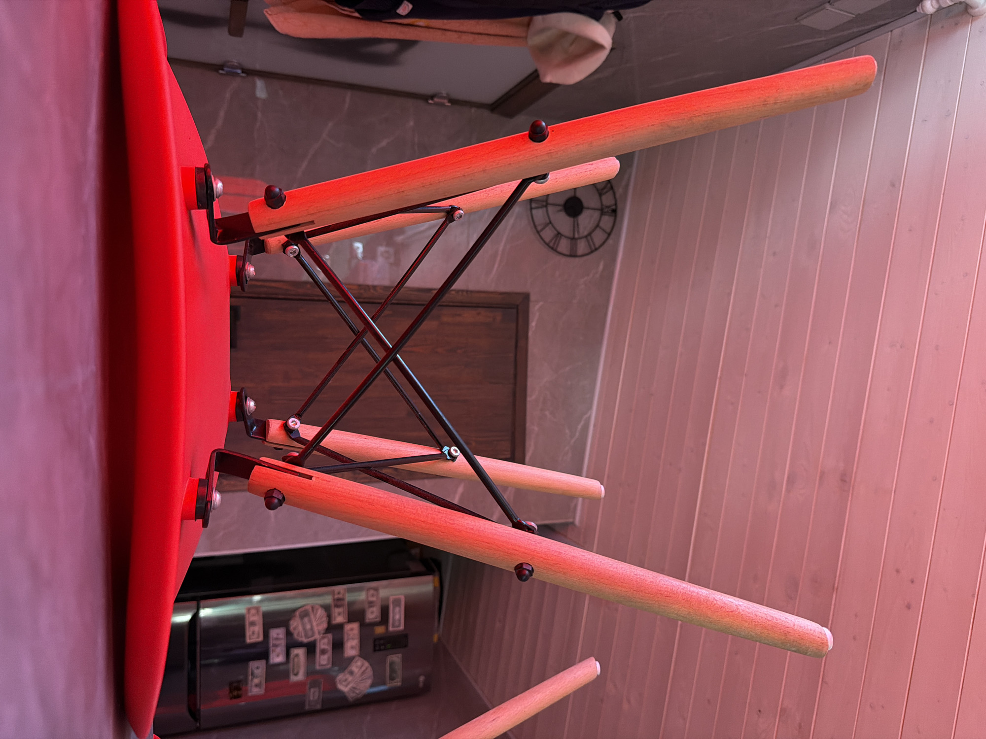The image shows a close-up, rotated view of a red chair's legs and seat. The chair is made of a red molded seat and wooden legs connected by black metal bracing. The background includes a wall with a wood-paneled texture and a marble-patterned surface, suggesting an indoor setting. A black clock with a round face is visible on the marble wall. Below the chair, a stainless steel appliance, possibly a refrigerator or dishwasher, is decorated with several square stickers. The lighting is warm and reddish, possibly from an artificial source, creating a pinkish hue across the scene. The orientation of the image is unusual, with the chair appearing to be upside down. No people are visible. The setting appears to be a room in a home or office in Orenburg, Russia.