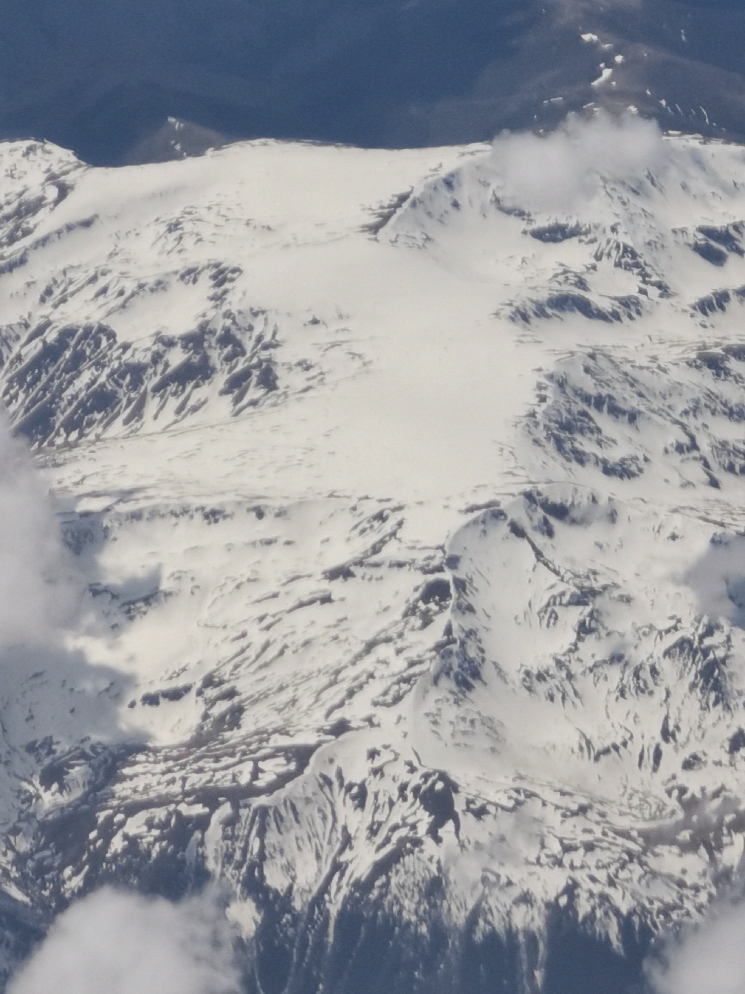 The image depicts a mountainous landscape covered in snow. The view is likely taken from an aerial perspective, possibly from an airplane. The rugged terrain is visible beneath the snow cover, revealing contours and ridges. Some cloud formations are present in the foreground, obscuring parts of the landscape. The image does not contain any recognizable elements specific to the city of Varna, Bulgaria. Therefore, it cannot be determined that the image was taken in Varna. There are no people, visible subjects or identifiable interactions in the image.