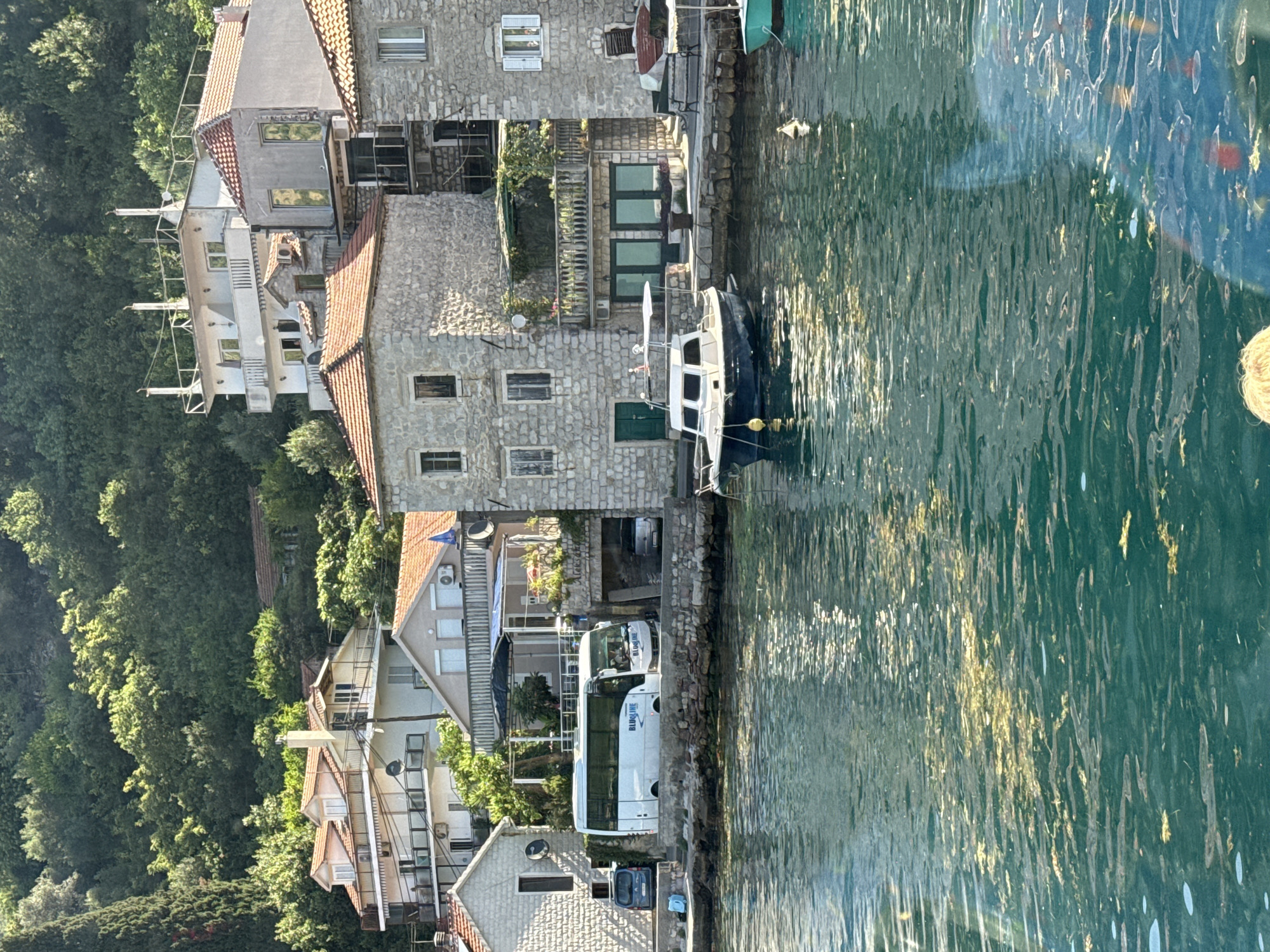 In the town of Lepetane, Montenegro, a scene unfolds along the waterfront. The clear, turquoise water of the bay is dappled with sunlight, reflecting the lush green hillside covered in trees that forms the backdrop. Along the shore, stone buildings with red-tiled roofs are nestled together.
In the foreground, a small, dark-hulled boat with a white cabin is moored in the water. Further along the quay, a white bus with "BLUELINE" written on its side is parked. Beside the bus, a small, dark-colored car is visible. Behind the bus, a modern white building with multiple balconies and large windows rises up the hillside.
The overall impression is one of a peaceful coastal settlement bathed in warm sunlight. The buildings display a mix of traditional stone construction and more contemporary architecture, indicating a blend of history and modernity in this picturesque location.