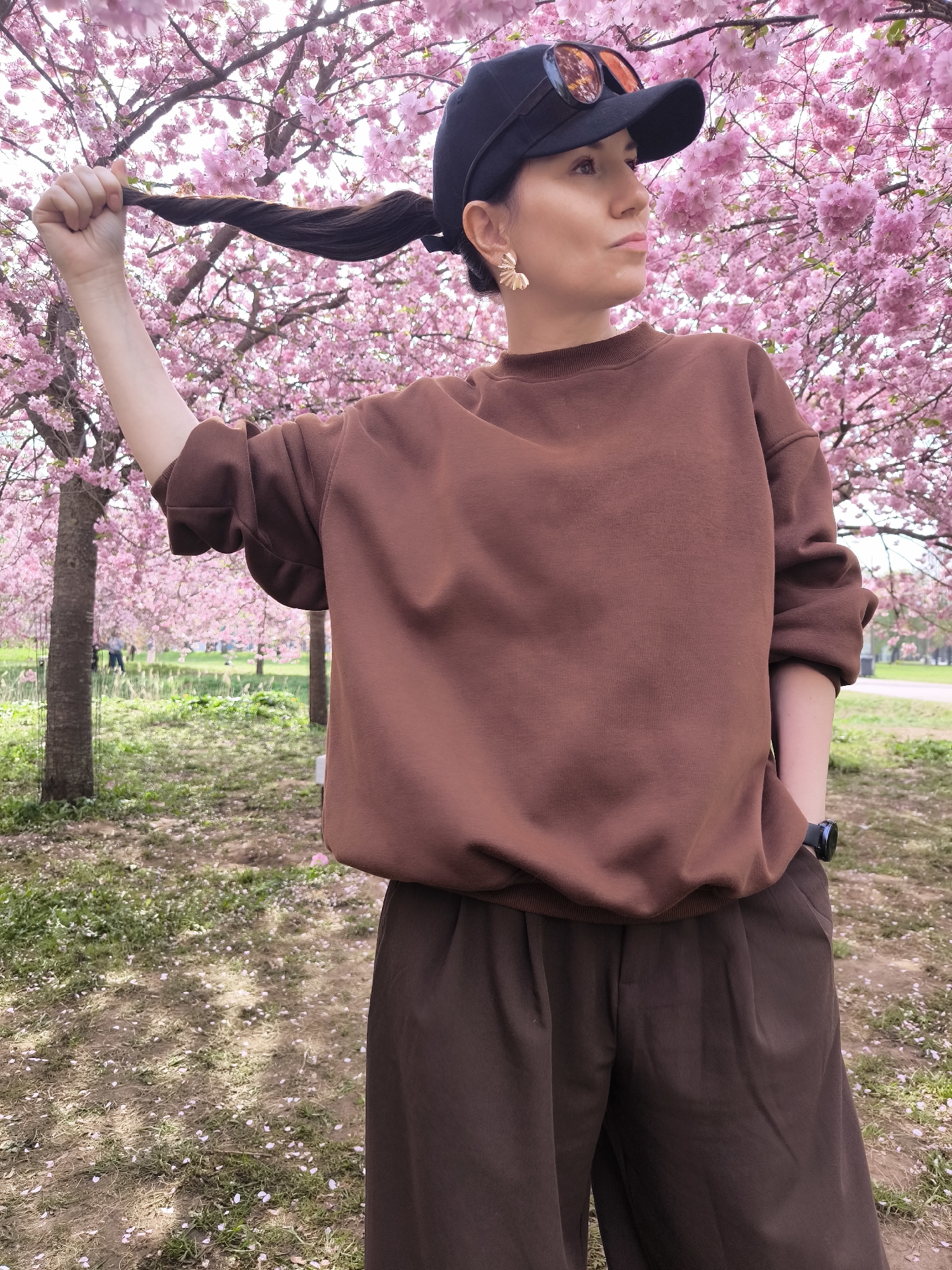 A woman stands in a park, with cherry blossom trees in full bloom forming the backdrop. She is wearing a brown sweater and matching brown pants, with a black baseball cap perched on her head and sunglasses atop the cap. The woman holds a strand of her hair in her raised left hand. Her right hand is casually placed in her pocket. She wears a watch on her left wrist and small gold-colored earrings. The park grounds are covered with grass and fallen cherry blossom petals. Other individuals are present in the background, further in the distance. The location is Riga, Latvia.