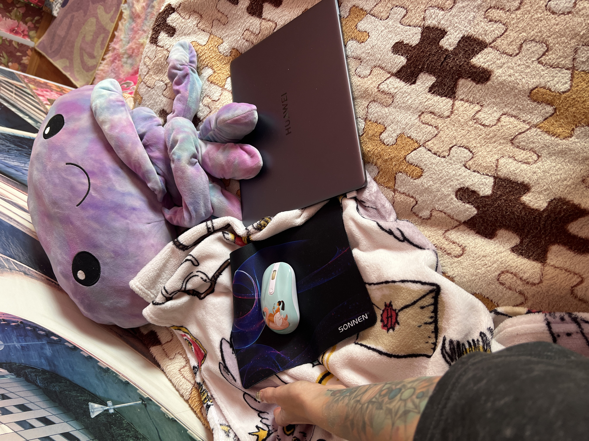 In Krasnoe-na-Volge, Russia, an image captures a close-up of a person's arm with tattoos resting on a plush blanket. A teal and white computer mouse with a cartoon character design sits on a black mousepad. Behind these items, a closed dark gray laptop with the "HUAWEI" logo is visible. The scene is further softened by a large, tie-dyed plush toy resembling a jellyfish draped over the laptop and extending onto the blanket. The blanket itself is adorned with a whimsical pattern of envelopes and flowers, and it lies on top of another textured blanket featuring a puzzle piece design in brown and beige hues. The background is out of focus but appears to show framed artwork on a wall.
