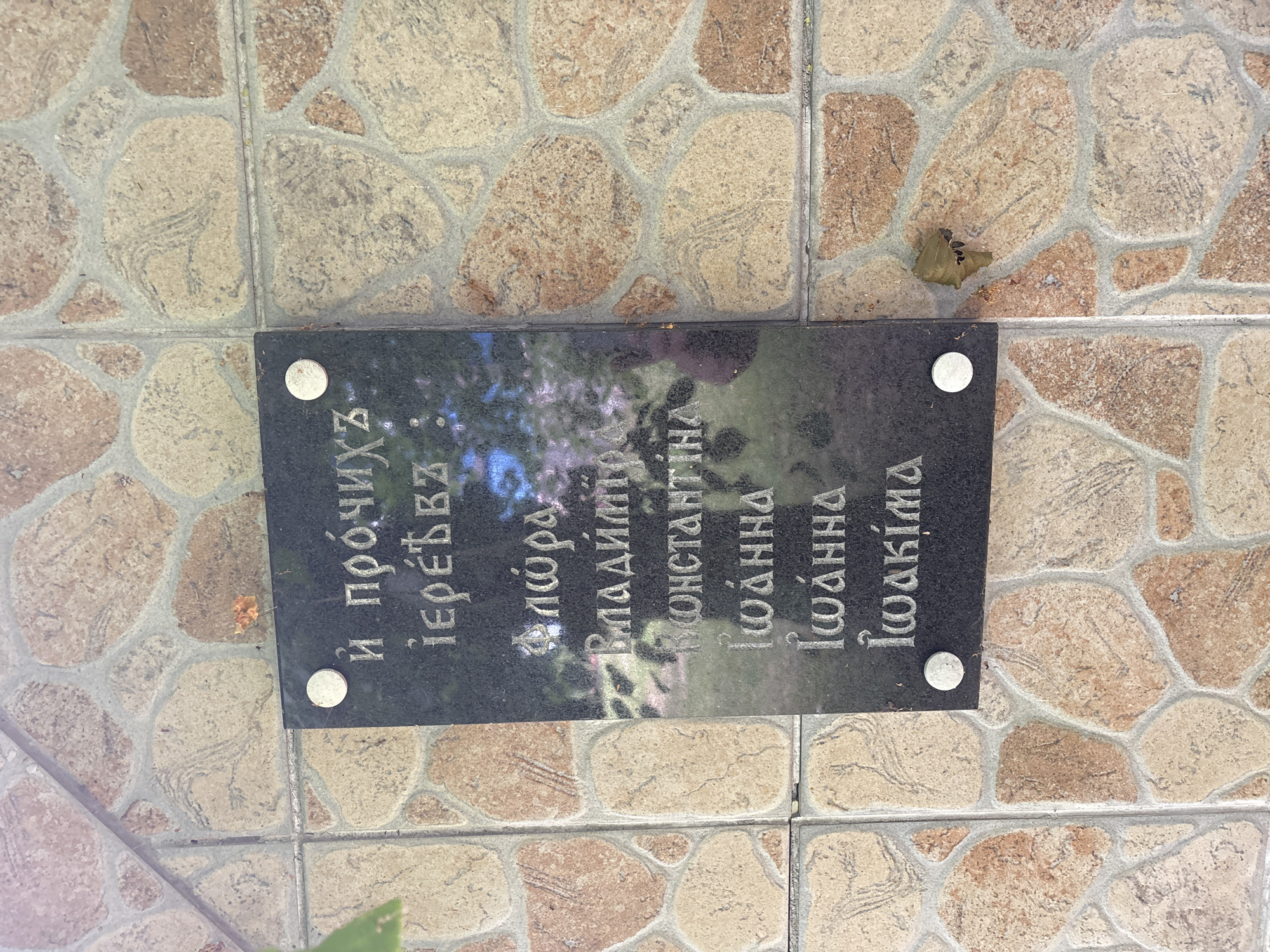 A black plaque with engraved text is affixed to a tiled wall. The text is in a Slavic script, likely Old Church Slavonic or a similar language. The plaque is mounted on a wall covered in irregularly shaped tiles in shades of beige, tan, and pinkish-brown, with grey grout. The lighting suggests it is daytime, with some dappled sunlight and shadows. A small, dried brown leaf is visible on the wall to the right of the plaque.
The text on the plaque appears to be a list of names or titles. The first line reads, "и прочиихъ". The second line reads, "іере́въ:". Subsequent lines appear to list names: "Флώра", "Владиміръ", "Константина", "Іωάνна", "Іωάνна", and "Іωакіма".
While the image itself does not provide direct information about the city of Puchły, Poland, similar plaques with such inscriptions are commonly found in Orthodox Christian churches or cemeteries. Puchły is known for its historical Orthodox church, the Church of the Transfiguration. Therefore, it is highly probable that this plaque is located at or near this church, serving as a memorial or record.