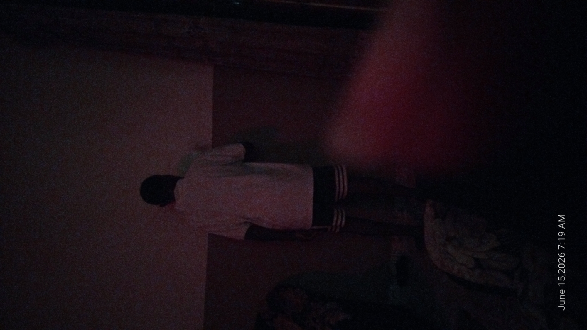 The image shows a person from behind, wearing a light-colored t-shirt and dark shorts with white stripes, standing in a dimly lit room. The person appears to be facing a wall. To the left of the person, a patterned fabric or blanket is visible. The room's walls are a muted reddish-brown. A bright, reddish-pink light source is visible in the lower right portion of the image, casting a glow on the wall and floor. The timestamp "June 15, 2026 7:19 AM" is overlaid at the bottom left of the image. The scene is consistent with an interior residential setting in Howayi, Nigeria.