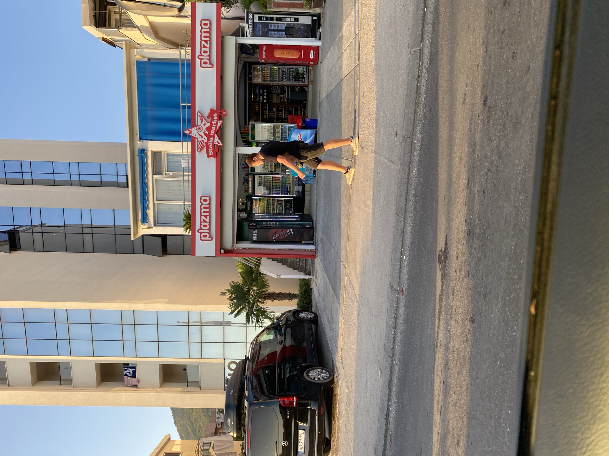 The image shows a street scene in Bar, Montenegro. In the foreground, a black Mercedes-Benz Vito van with a roof box is parked. A man in a black t-shirt and shorts is walking on the sidewalk in front of a "Plazma" convenience store. The store has a bright red and white awning and features several refrigerated display cases stocked with beverages. To the left of the store is a tall, modern building with a glass facade. The scene is illuminated by the warm light of the setting sun.