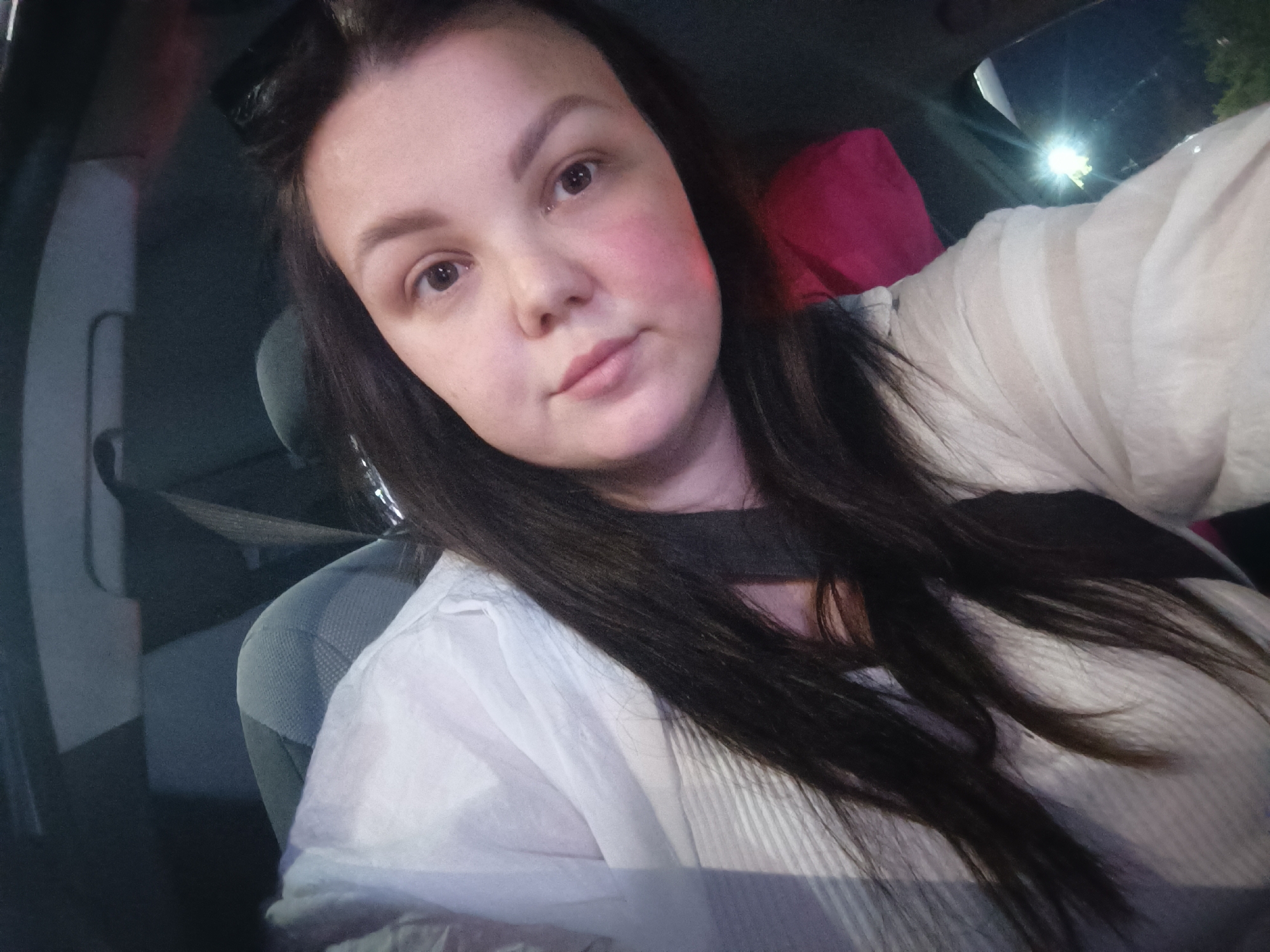 A woman with fair skin and dark hair is seated in a car, likely the driver's seat. She's wearing a white shirt and a dark-colored top underneath. The car's interior is visible, including the headrest and seat upholstery, suggesting an ordinary vehicle. Outside, a light source is present, possibly a streetlight, indicating it's either evening or night. The exact location within Chaikovsky, Russia, is impossible to determine from the image alone.
