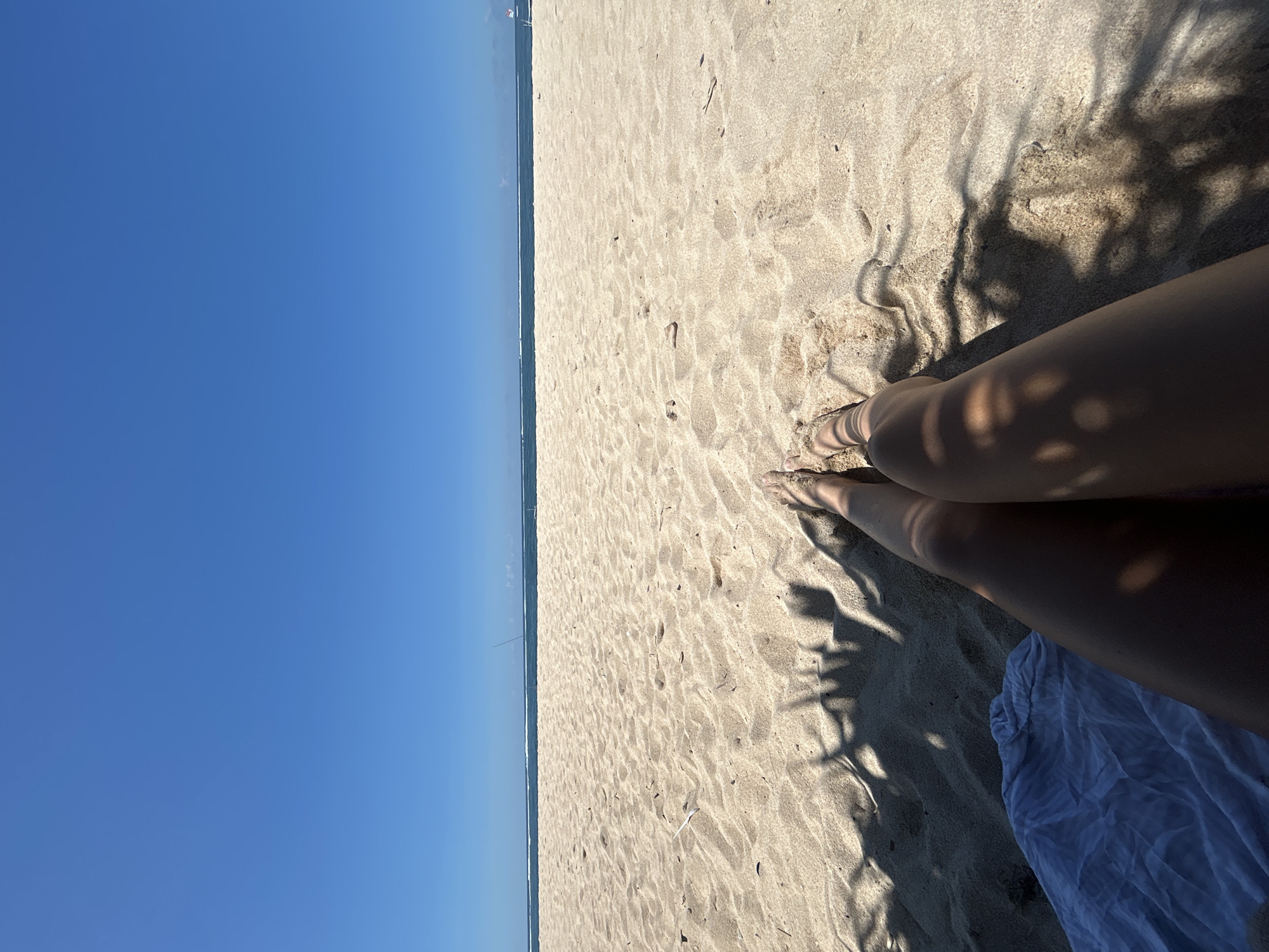 A beach scene in Kuta Selatan, Indonesia, captures a sunny day. The horizon line, where the ocean meets the clear blue sky, runs along the left side of the frame. The sandy beach extends across the right side, marked with footprints and shadows, including the shadows of tree foliage and what appear to be human legs. A white cloth rests near the legs. The photo is taken from a high angle, possibly from a person sitting on the beach.