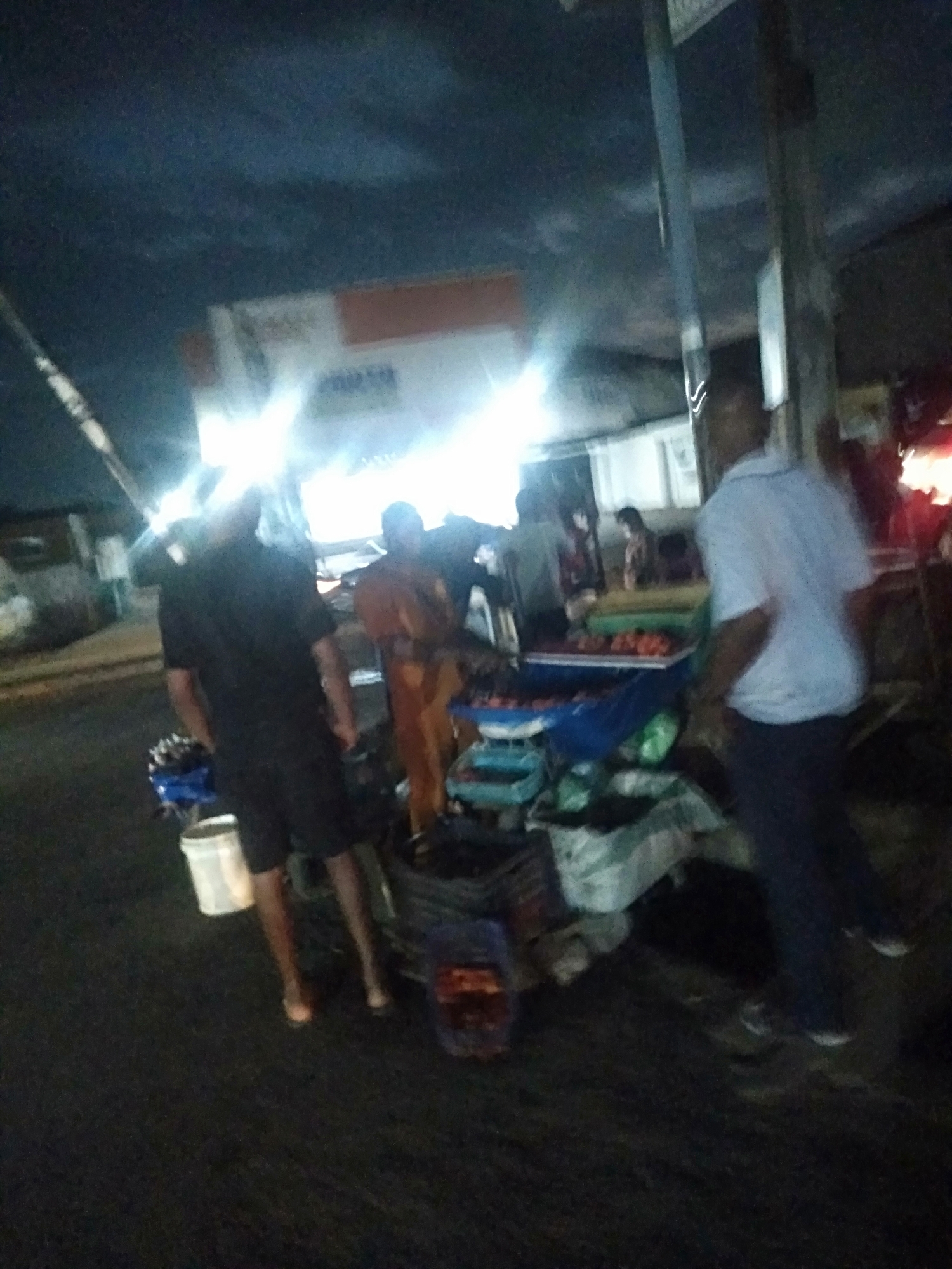 The image shows an outdoor night scene, presumably in Shangisha, Nigeria, given the location reference. Several individuals are clustered around what appears to be a street vendor's setup. The vendor's table displays fruits and produce. A man in a white shirt and dark pants stands to the right. A man in black clothing is standing next to a white bucket. There's another man in an orange-colored garment standing by the vendor's table. The background features a building, with a bright light source shining from it. The overall lighting of the image is poor.