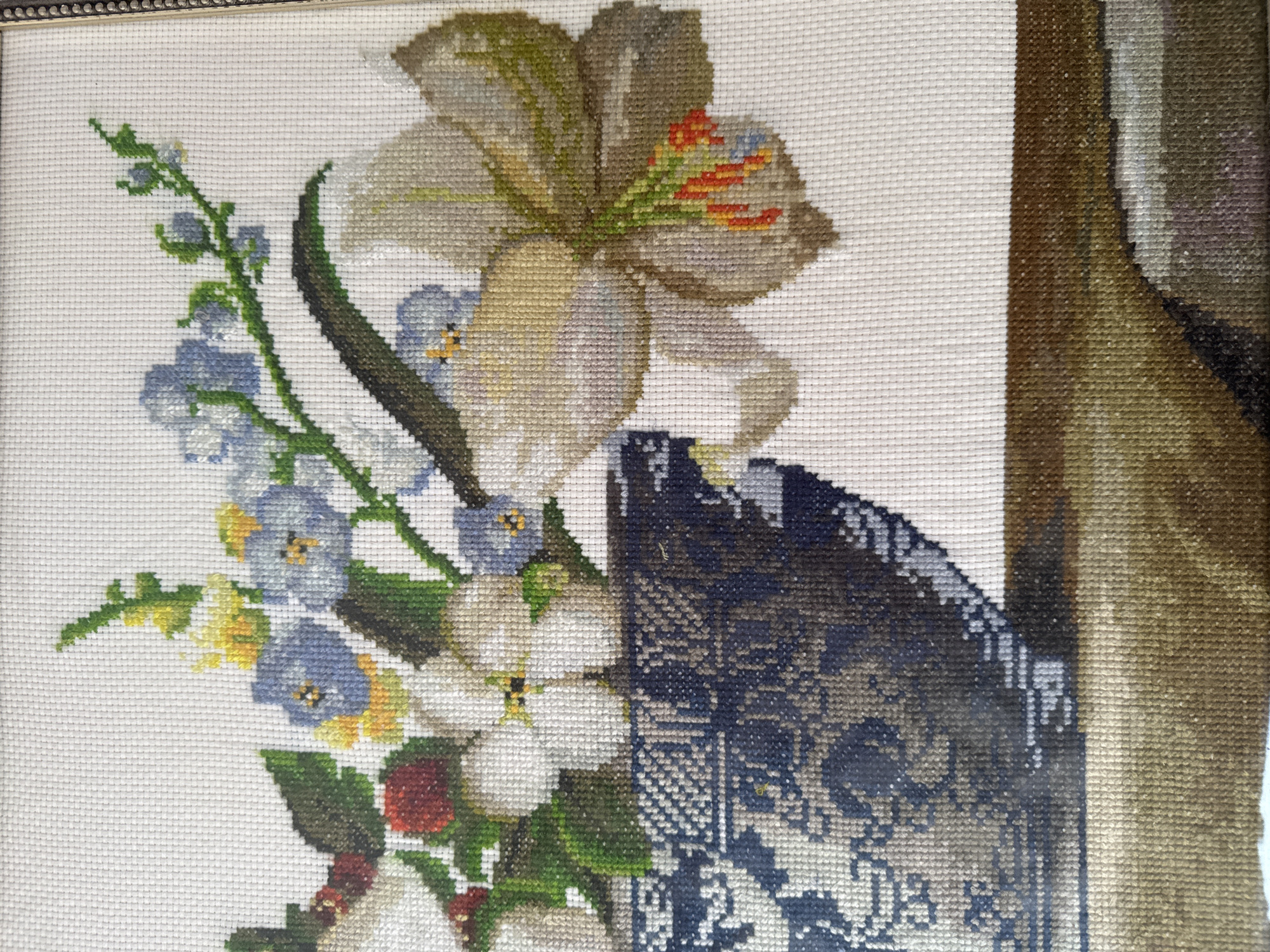 This is a close-up, eye-level, medium shot of a framed cross-stitch embroidery of a floral arrangement. The embroidery depicts a bouquet of white, blue, and yellow flowers with green stems and leaves, as well as some red berries. The bouquet is placed in a dark blue and white patterned vase, which sits on a brown surface. The background is a plain white canvas. The cross-stitch technique is evident in the grid-like pattern of the threads. There are no people or animals visible in the image. The lighting is even and natural, suggesting an indoor setting. The composition is centered on the floral arrangement and vase. The image does not provide any information about its location in Bishkek City, Kyrgyzstan.