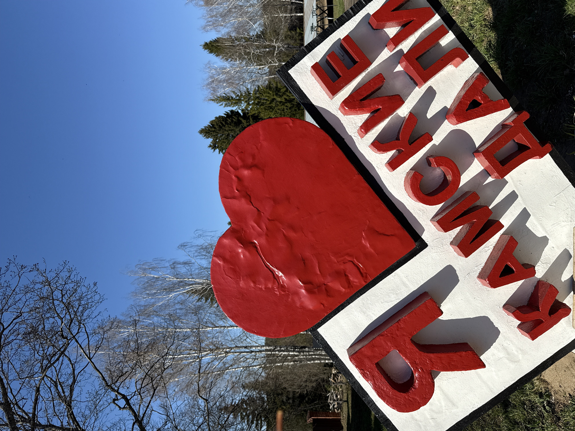 The image shows an outdoor sign in Galevo, Russia, featuring a large red heart and red Cyrillic letters on a white background. The sign appears to be located in a park or green space, as trees and grass are visible in the background. The sky is clear and blue.