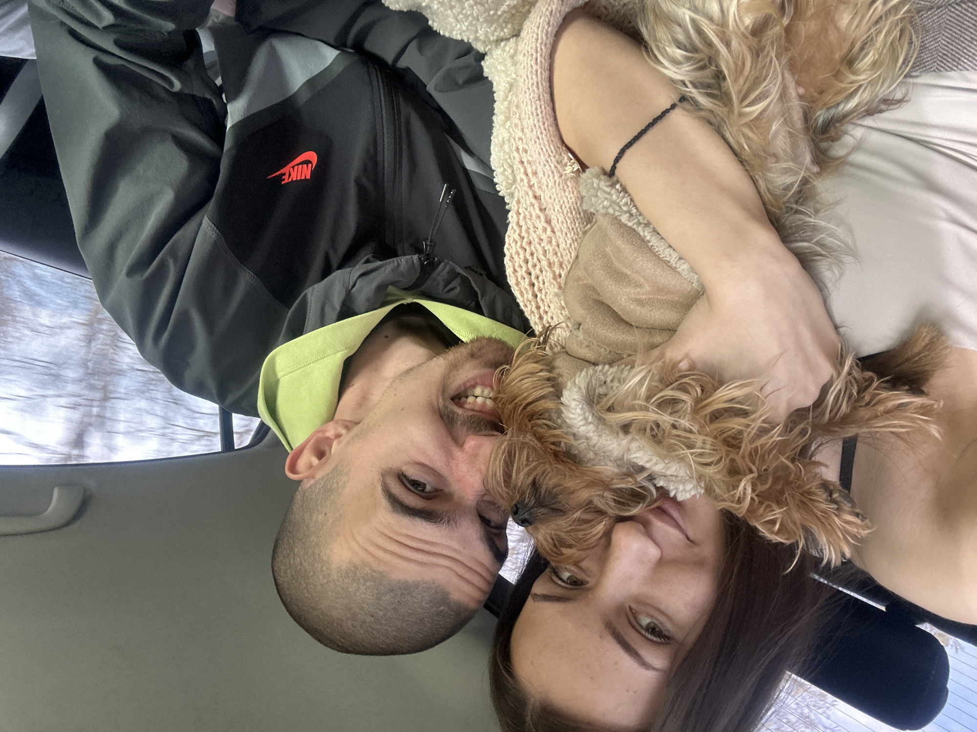 A selfie of a man and a woman, along with a dog, taken inside a car. The man, wearing a black Nike jacket with a green hoodie underneath, is on the left and the woman is on the right. The dog is between them, nestled against the woman. The background shows a view out the car window, appearing to be winter. The car interior is visible. The location is not discernable to be Marinka, Bulgaria based on the image.