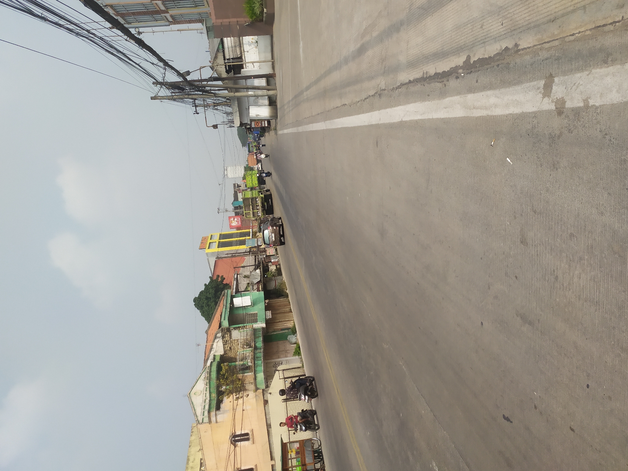 The image depicts a street scene in Kragilan, Indonesia, on a bright, overcast day. The street is paved with asphalt and has a white dividing line. On the left side of the street, there are several buildings, including a yellow building with a sign and a brick building with multiple stories. Some of these buildings appear to be businesses or shops, with some showing signs of wear. There are also several vehicles visible, including two motorcycles with riders, two trucks, and a car. The motorcycles are on the road, with one rider wearing a red shirt and the other wearing a darker shirt. The trucks are also on the road, with one being green and the other green and yellow. The car is brown. A food cart with a green canopy is parked on the side of the road. The right side of the street has a sidewalk and more buildings, including a brick building with windows. There are numerous electrical wires and poles lining the street. The sky is mostly clear with some scattered clouds. The overall impression is that of a moderately busy urban street.