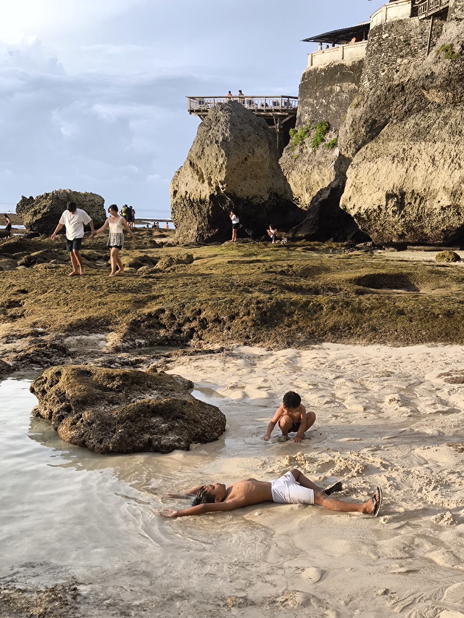 The photograph captures a beach scene in Uluwatu, Indonesia, depicting a mix of leisure and interaction against a backdrop of natural rock formations.
In the foreground, a person lies prone on the sand, partially submerged in shallow water. They are wearing white shorts and sandals, with their arms extended. Nearby, a child is crouched on the sand, appearing to be playing. A large, dark rock sits partially submerged in the water, close to the recumbent person.
In the middle ground, a couple is walking on a mossy, rock-strewn area. Behind them, other people are visible, some standing and others interacting near the rocks.
In the background, a cliff-like rock formation rises, topped with a building that seems to be a restaurant or viewing platform, with people visible on the structure. The sky is overcast with light clouds. The general setting evokes a coastal environment with exposed rock formations and shallow tide pools.