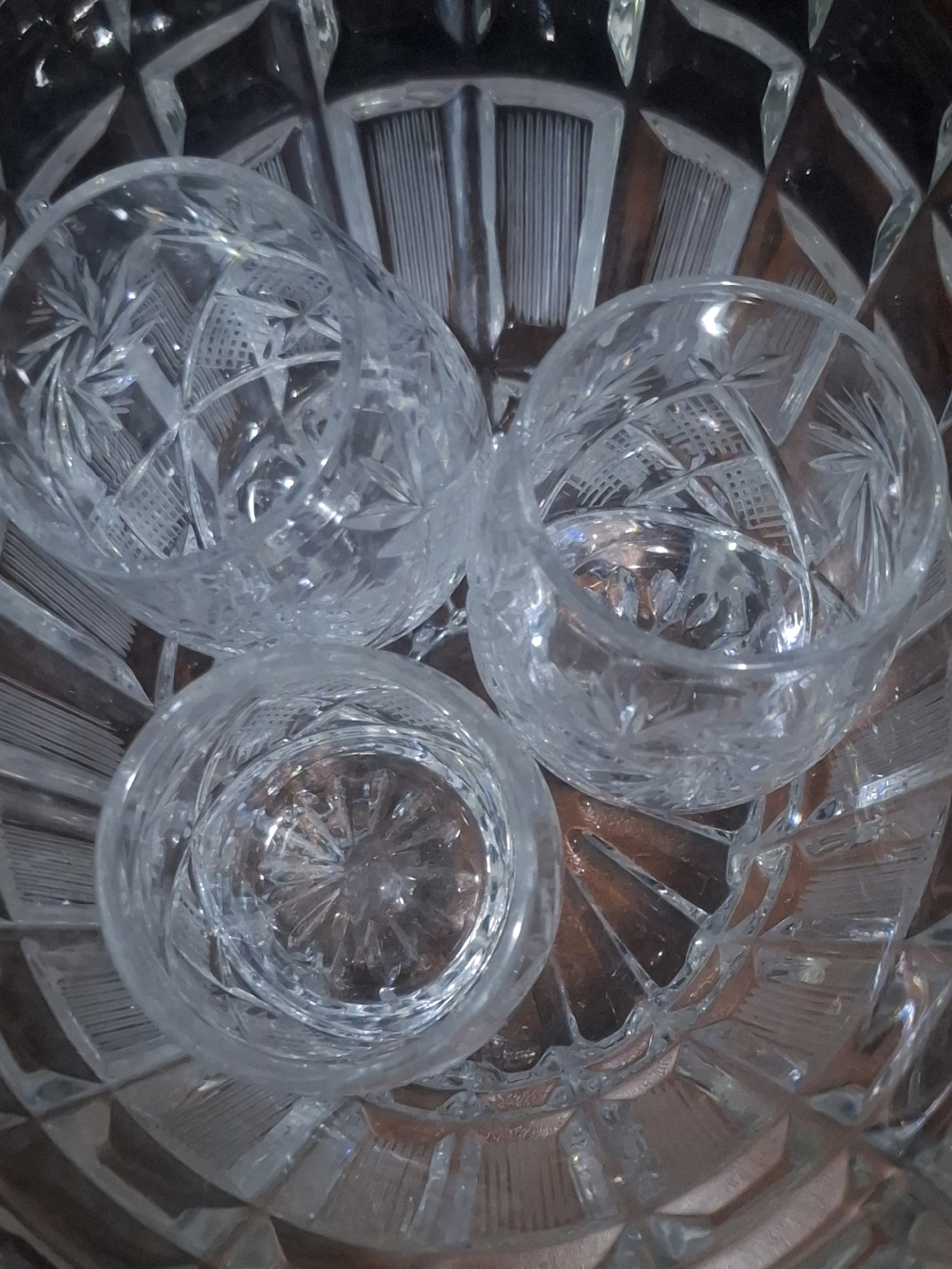 The image is a close-up, overhead shot of three crystal glasses arranged in a larger crystal bowl. The glasses have intricate cut patterns that refract light, creating a shimmering effect. The bowl itself also features a decorative cut design. The lighting suggests an indoor environment. There are no people visible in the image. The location in Almaty, Kazakhstan, is not discernible from the visual content of the image.