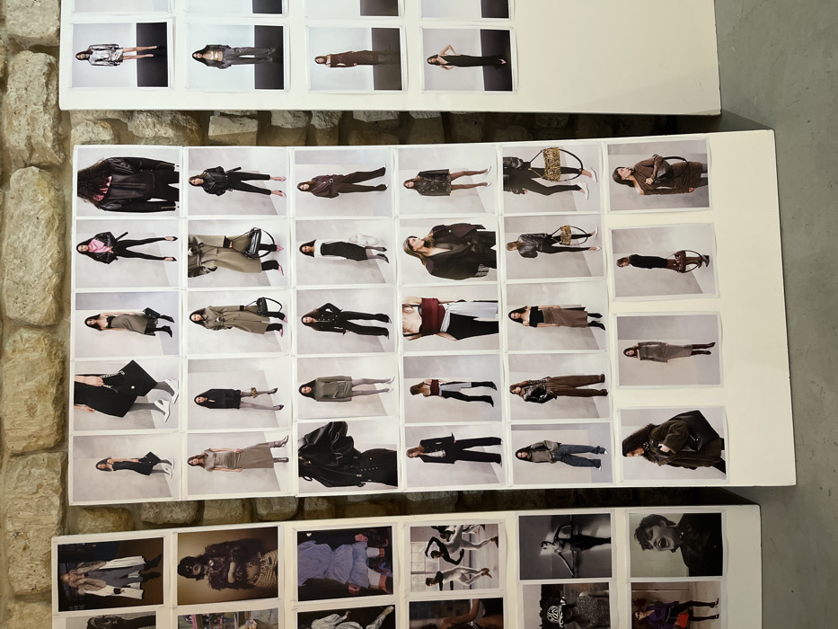 The image displays an interior space within Paris, France, featuring three freestanding white display boards against a rough, light-colored stone wall. The floor is a dark grey concrete. The boards are covered with numerous printed photographs, primarily arranged in grids.
The central and largest board presents a densely packed grid of approximately 35-40 vertical, color photographs. Each photo depicts a female model in a distinct fashion ensemble, typically against a plain, light studio background. The models showcase a variety of garments, including coats, jackets (leather), dresses, skirts, trousers, knitwear, and accessories like handbags and scarves. Poses are static and professional, focused on presenting the clothing. Colors observed are largely muted, featuring blacks, greys, browns, olives, and occasional pops of color like pink and maroon.
To the left, a narrower white board displays fewer, larger photographs. These include images of a woman in a long coat, a person with intricate face paint or a mask, a light blue ruffled dress, and several black-and-white photos of figures in dynamic, possibly dance-related, poses. The bottom left shows a woman in a purple outfit and a close-up of a person with short hair.
A third, partially visible white board on the far right also features grids of fashion photographs, similar in style to the central board, showing models in items like a metallic silver dress and long dark dresses.
The scene conveys a fashion-related presentation, likely a collection mood board, lookbook display, or backstage styling reference, in a functional, minimalist setting. No individuals are present within the primary scene beyond the photographed models.