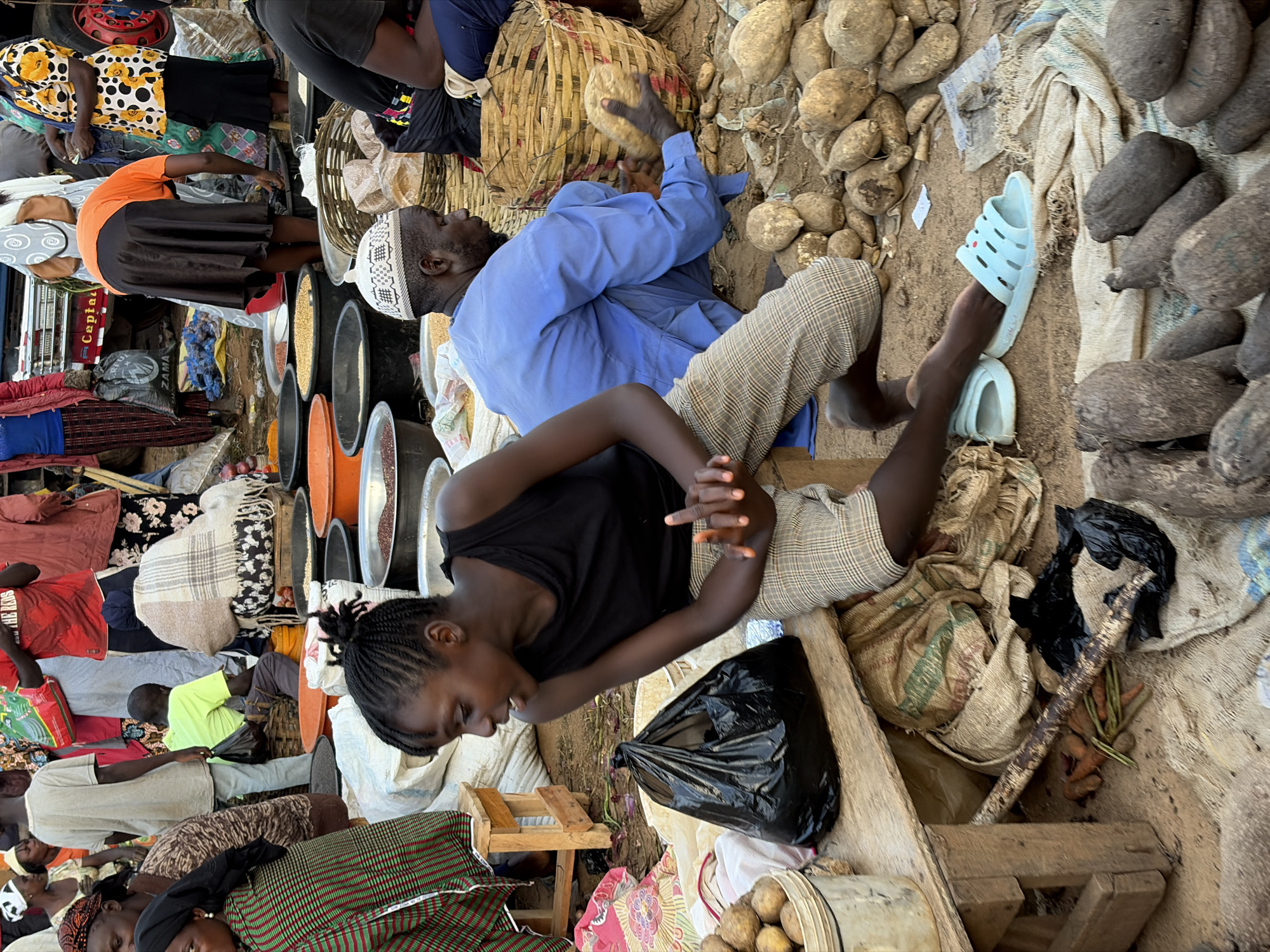 This is a medium shot of a bustling outdoor market scene in Gura Top, Nigeria. A young woman with braided hair, wearing a black tank top and checkered pants, is seated and looking to her left, her right hand raised and fingers splayed. Beside her, on a wooden bench, rests a large black plastic bag and a bucket filled with what appear to be potatoes. In front of the woman, a light blue sandal is visible on the ground.
To her right, a man wearing a light blue traditional robe and a white and black patterned cap is kneeling, reaching into a woven basket. He holds a large root vegetable, possibly a yam, in his hands. The ground around him is covered with dirt, sacks, and numerous similar root vegetables. In the background, various individuals are standing and milling about, engaged in market activities. There are large containers filled with grains and other market goods, along with more sacks and baskets. The overall environment suggests a vibrant, active marketplace.